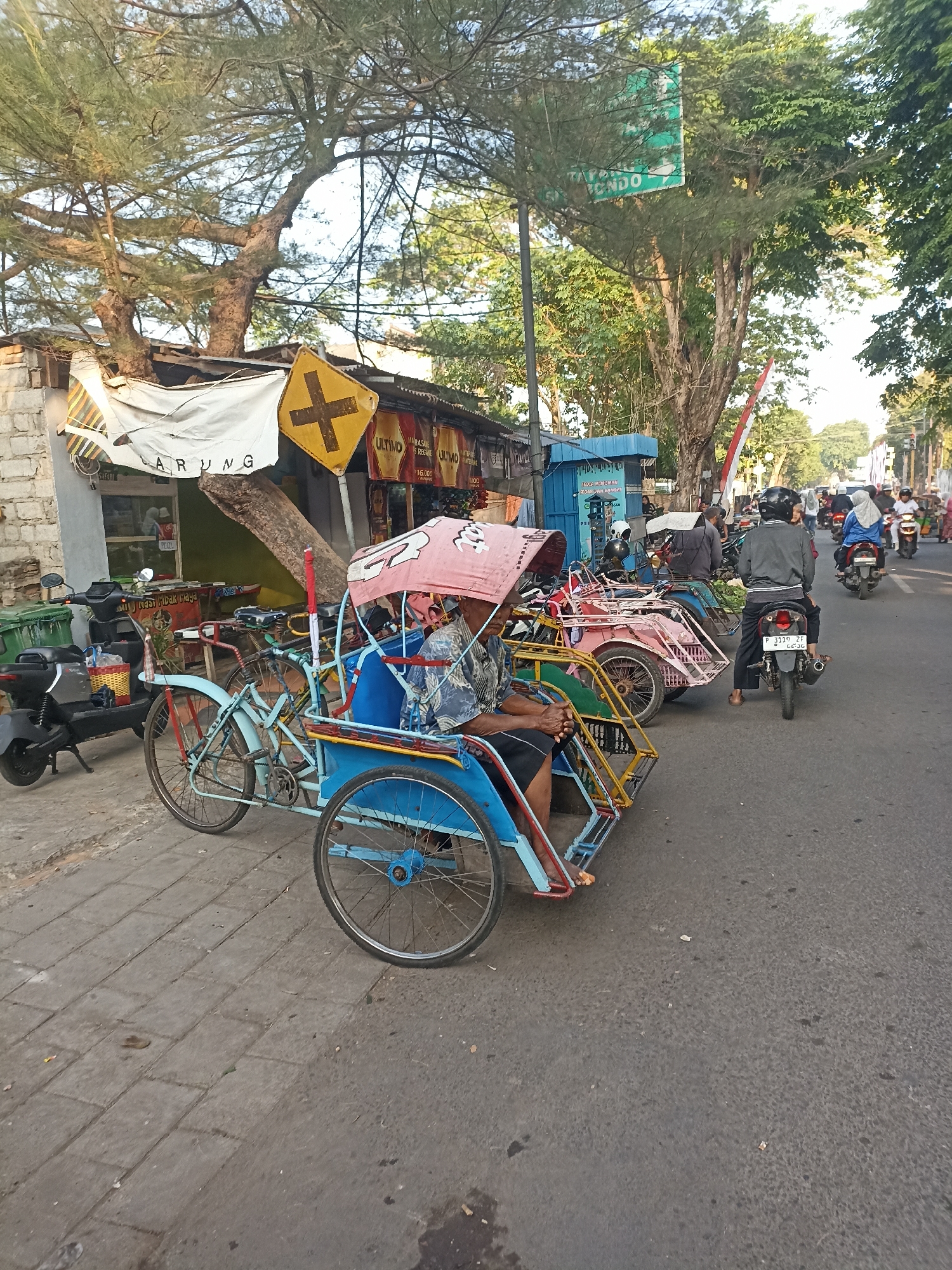 The image depicts a street scene in Kelurahan Kepatihan, Indonesia, with a focus on a row of traditional pedicabs parked on the side of a road. The pedicabs, also known as becak, are brightly colored with a pink canopy on the leading one. A man is seated in the driver's position of the foremost pedicab. Behind the pedicabs, there are small shops or stalls with various goods displayed, including what appears to be fabric or clothing. The shops have awnings and signs, one of which has the word "ARUNG" visible. Several trees line the street, with their branches casting shadows. A road runs alongside the sidewalk, with several motorcycles and pedestrians visible. A green trash bin is positioned near a parked black scooter. A yellow road sign with a black cross symbol is mounted on a pole near the shops. The overall atmosphere suggests a bustling local market or street commerce area, typical of Indonesian urban environments. The time of day appears to be either early morning or late afternoon due to the low angle of the sunlight.