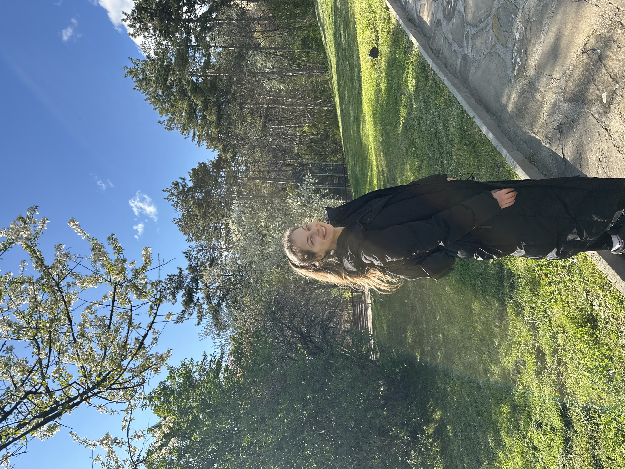 A young woman with long, light brown hair stands outside in a park-like setting. She is wearing a black jacket. The background features a lush green lawn, trees, and a blue sky with some clouds. There is a stone path visible to the right of the frame. The location is Stara Zagora, Bulgaria.
