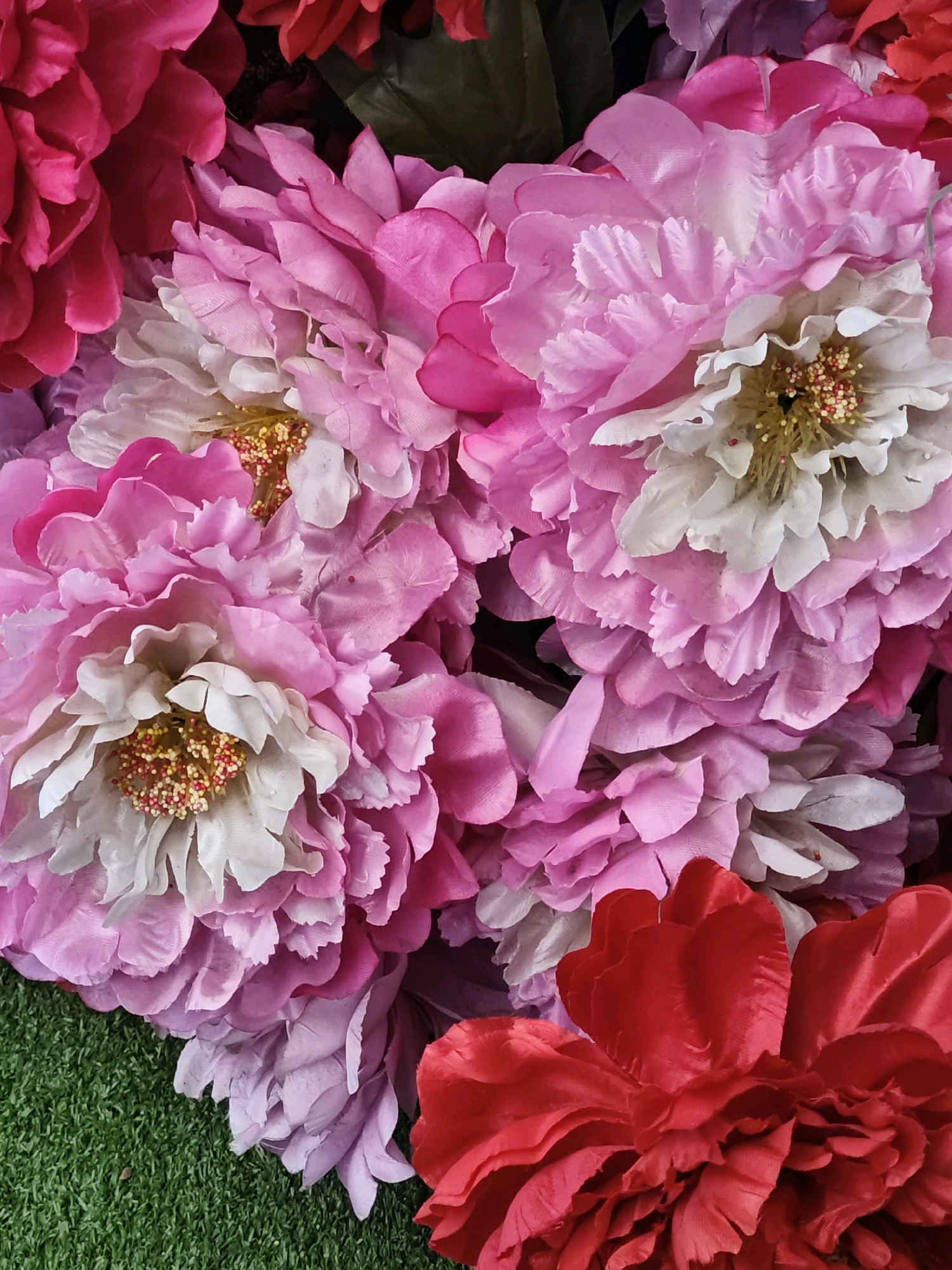 This is a close-up, top-down, studio shot of a vibrant, artificial flower arrangement with a predominantly pink and red color palette. The flowers appear to be peonies or similar large, multi-petaled blooms. Some flowers are a bright fuchsia, while others are a softer pink, and a few have white petals at their center. The centers of the white-petaled flowers are adorned with yellow stamens tipped with red. A few deep red flowers are also visible, one prominently in the lower right corner. The arrangement is set against a backdrop of artificial green grass, which is visible in the lower left quadrant of the image. There are no people, animals, or any discernible landmarks that would indicate a specific location in Moscow, Russia, or any other city. The focus is entirely on the texture and color of the floral display.
