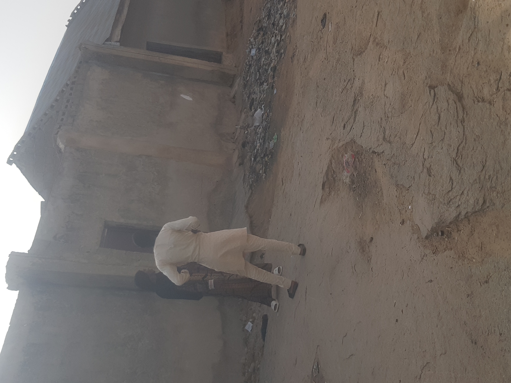 In the Dabawa, Nigeria, an outdoor scene features a weathered, gray concrete building on the left side of the frame. Debris and refuse are scattered along the base of the building and extend onto the dusty, cracked ground. Two individuals are present: one wearing a light-colored, possibly white, traditional outfit, and another individual wearing a dark colored outfit are positioned close to the building; only the feet and lower legs of the second person are visible. The scene is illuminated with a diffused, natural light. The ground surface has a coarse texture with visible cracks and variations in color.