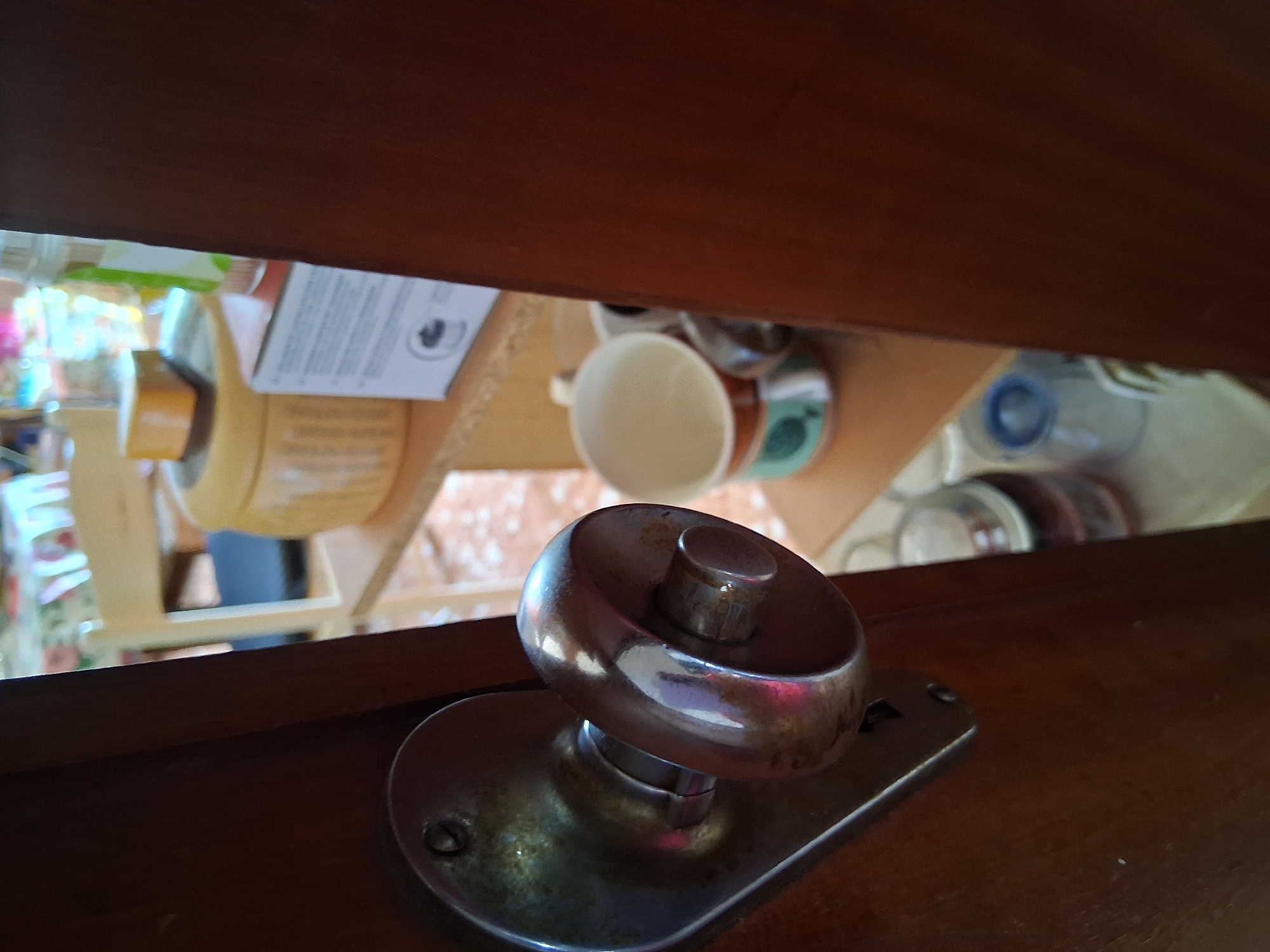 The image shows a close-up of a vintage metal doorknob on a wooden door. The doorknob is round and has a reflective surface with some signs of age and wear. It is mounted on a decorative metal plate with a screw visible. The door appears to be partially open, revealing a glimpse of a room in the background.
The background is out of focus but suggests a domestic or commercial setting. There are shelves with various items, including a cylindrical container, a box, and several mugs or cups. A wooden chair is also partially visible. The lighting is somewhat dim, with reflections on the doorknob indicating a light source.
The scene is likely located within the city of Colorno, Italy. The style of the doorknob and the general ambiance suggest an older building, possibly a historical residence or a traditional shop. The objects on the shelves might indicate a kitchen, pantry, or a display area. There are no people visible in the image.