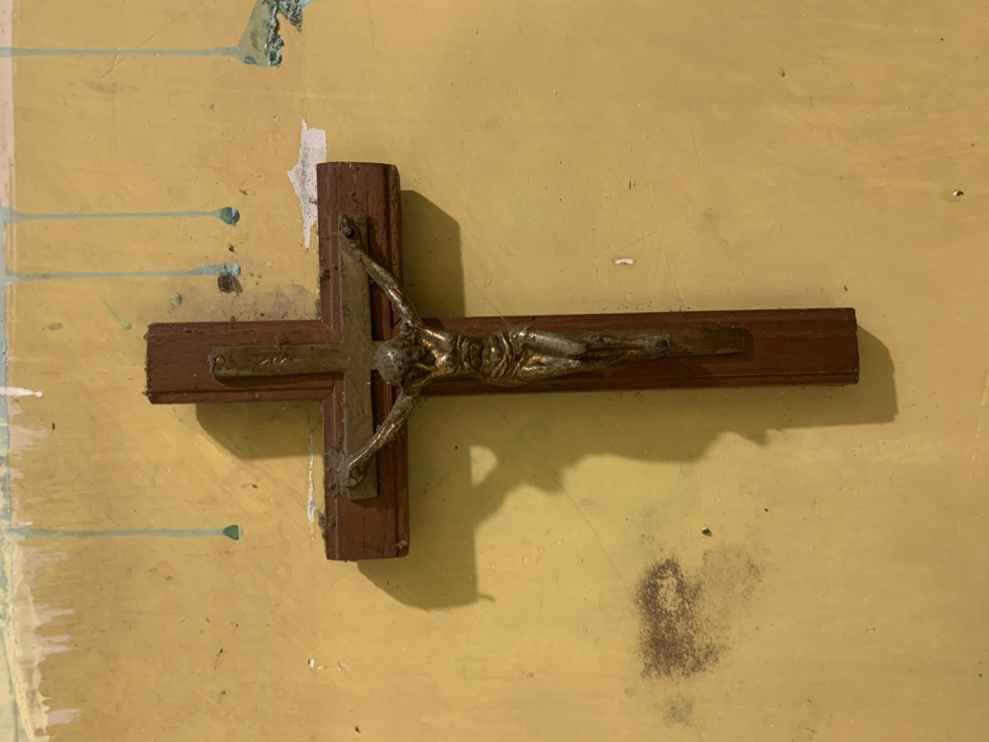 A wooden crucifix with a brass figure of Jesus Christ is mounted on a textured, yellow-painted wall in Dukuh Mojo, Indonesia. The wall shows signs of age and wear, with streaks of light blue paint dripping from the top and some areas of discoloration, including a dark, irregular stain to the left of the crucifix. The lighting casts a distinct shadow of the crucifix onto the wall.