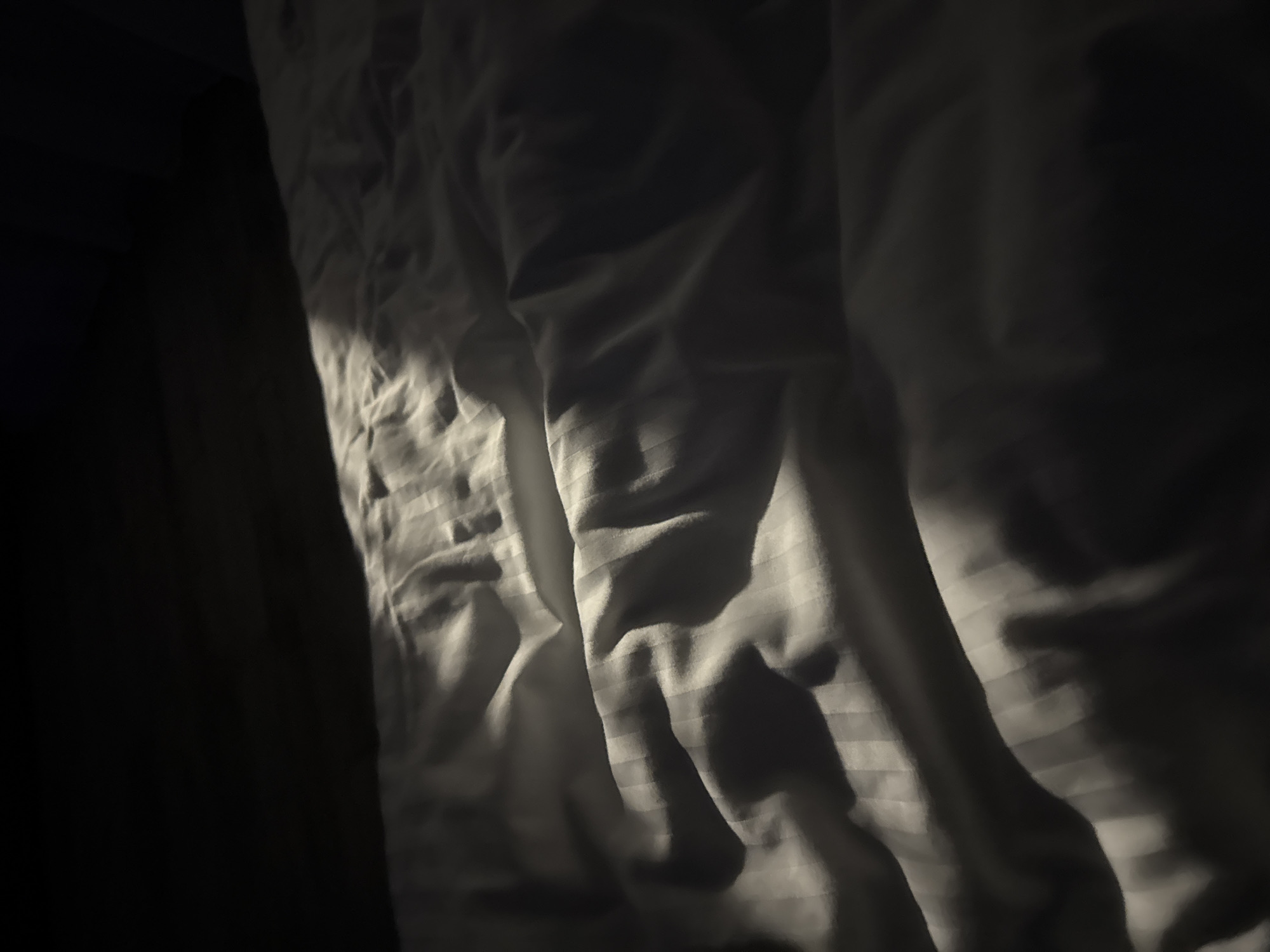 The image displays a close-up, low-angle view of a rumpled white bed with a subtle striped pattern. A diagonal shaft of light cuts across the bedding from the upper left, creating dramatic shadows and highlighting the texture of the fabric. The upper portion of the image is very dark, obscuring any details of the environment. The scene appears to be indoors, possibly a bedroom. There are no people or specific landmarks visible that would indicate a location in Doma 1096 Km, Russia.
