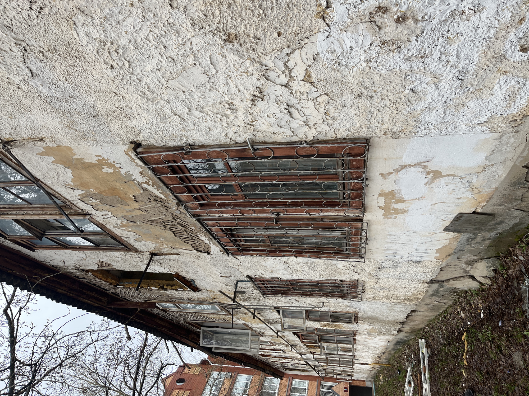 An upward-angled outdoor shot shows a decaying building facade in what appears to be Chaikovsky, Russia. The facade is white with rough textured plaster and patches where the plaster has peeled off, revealing the material underneath. Windows with barred covers are spaced along the facade. Some windows have visible panes, while others are dark or obscured. Beneath the facade is the visible roofline. Below and to the left of the roofline are bare trees with thin branches. The bottom of the frame shows a partial view of another building with red brick and white-framed windows. The right edge of the frame shows debris and possible vegetation. The overall condition of the building suggests neglect or disrepair.