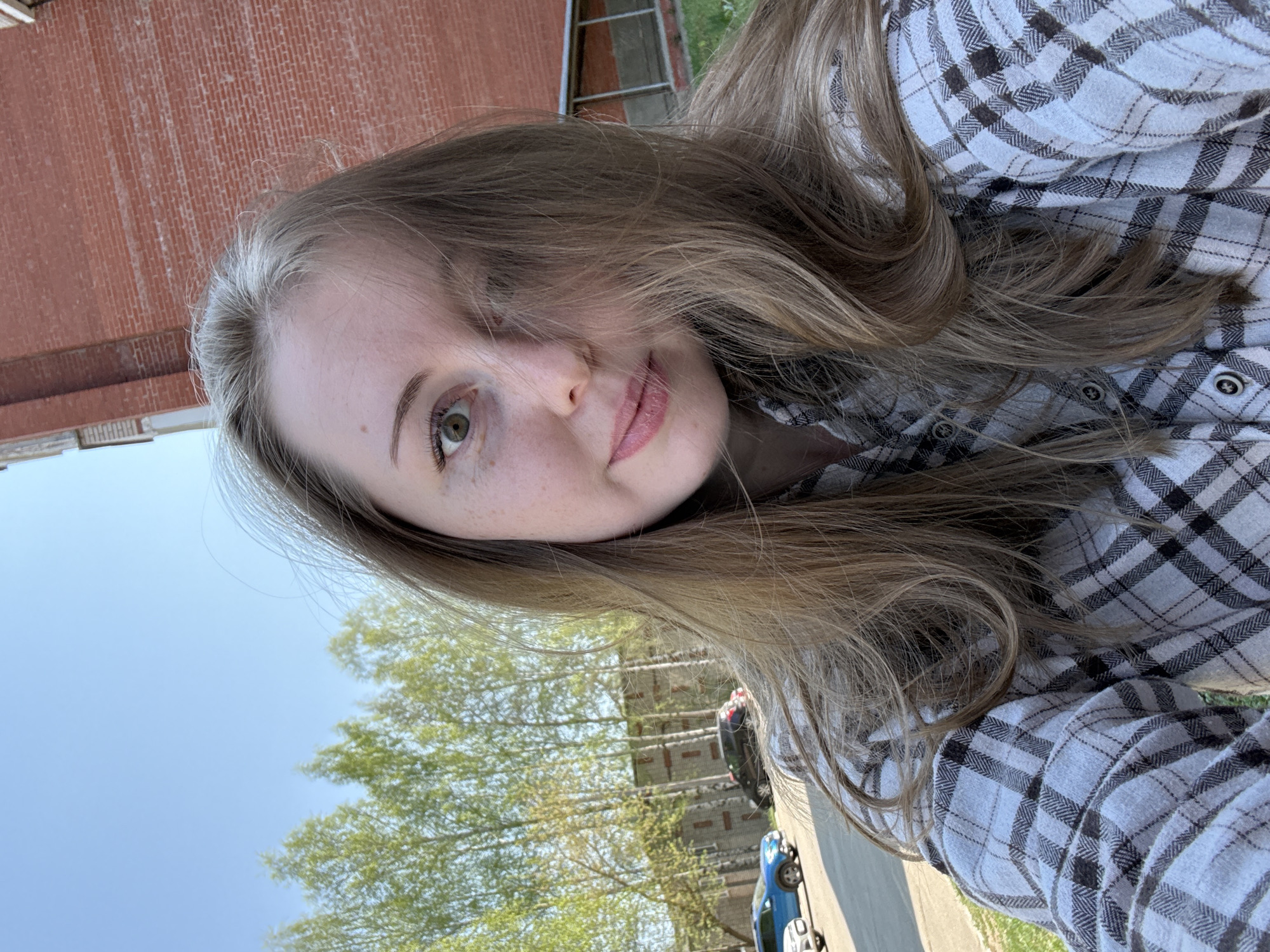 A woman with light brown hair and fair skin is looking at the camera. She is wearing a gray and white plaid shirt. Behind her is a brick building and a row of trees. The sky is blue. Two parked cars can be seen in the background. The location is an urban area, with a building, street, trees and sky visible.