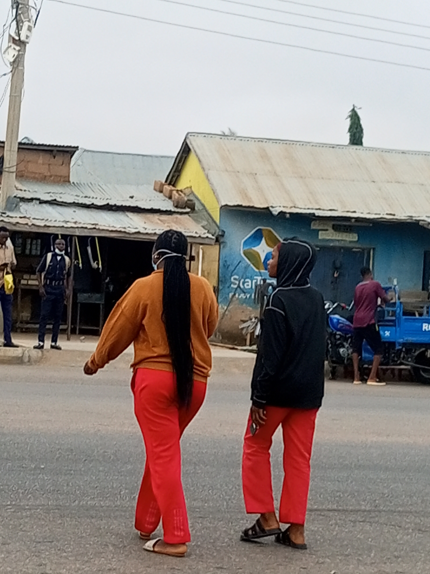 This image, taken in Tumfure, Nigeria, depicts a street scene with several individuals and buildings. In the foreground, two women are walking away from the camera on a paved road. The woman on the left wears an orange sweater and red pants, with her long black hair styled in braids. The woman on the right is dressed in a black hooded jacket and red pants.
In the background, a building with a corrugated metal roof and a yellow facade is visible, appearing to be a shop or commercial establishment. A man in a blue uniform with a mask is standing near the entrance of this building. To the right, another man in a burgundy shirt and dark shorts is seen near a blue three-wheeled vehicle. A motorcycle is parked behind him. The sky is overcast, and utility poles with wires are present on the left side of the image. The overall environment suggests a moderately busy urban or semi-urban area.