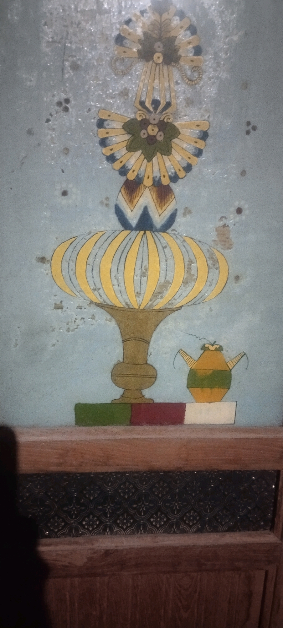 The image shows a close-up of a faded mural painted on a light blue, textured wall. The mural depicts a decorative, symmetrical floral or fan-like motif at the top, with colors of gold, dark blue, and brown. Below this is a stylized vase or urn shape with yellow and light blue stripes, resting on a golden pedestal. To the right of the pedestal, a small, green and yellow teapot-like object is depicted. At the bottom of the mural, there are three rectangular blocks of color: green, red, and white. The mural is positioned above a wooden surface with a dark, patterned glass panel. The overall condition of the mural suggests age and wear. The location in Daleman, Indonesia, is not visually represented in the image itself, but is provided as context.