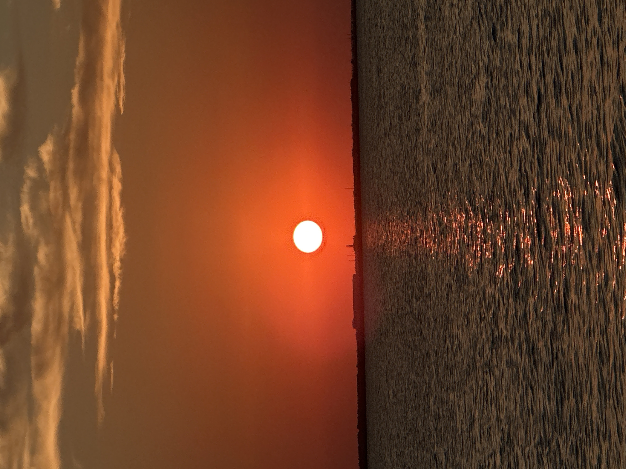 This is a photograph of a sunset over a body of water, likely a lake or sea. The sun is a bright orange orb centered in the frame, casting a warm glow across the sky. Wispy, orange-tinted clouds stretch across the upper portion of the image. A dark, horizontal line separates the sky from the water, representing the distant shoreline with some faint structures visible. The water in the foreground is dark and rippling, reflecting the orange hues of the sunset, creating a shimmering path of light leading towards the viewer. The overall mood is serene and peaceful. Without further information, the specific location in Kokshetau, Kazakhstan cannot be determined from the image alone.