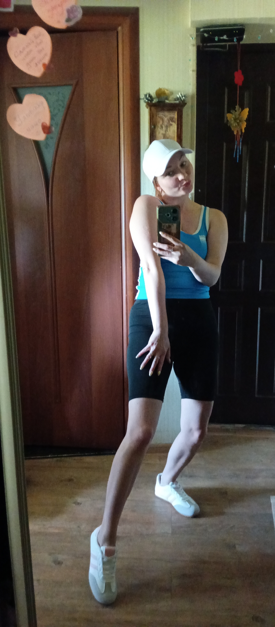 A fair-skinned woman stands in front of a mirror, holding a phone to take a selfie. She is wearing a white baseball cap, a blue tank top with white straps, black athletic shorts, and white and gray sneakers. Her hair is light brown or red. Her nails are long and painted a light color. She is striking a pose with one leg forward and her hand resting on her thigh. Her mouth is pursed in a kiss.
The location is an interior space with a hardwood floor and a brown wooden door with heart-shaped decorations on it. A dark wooden door is visible in the background. A wall clock with decorative elements is hanging near the dark wooden door.