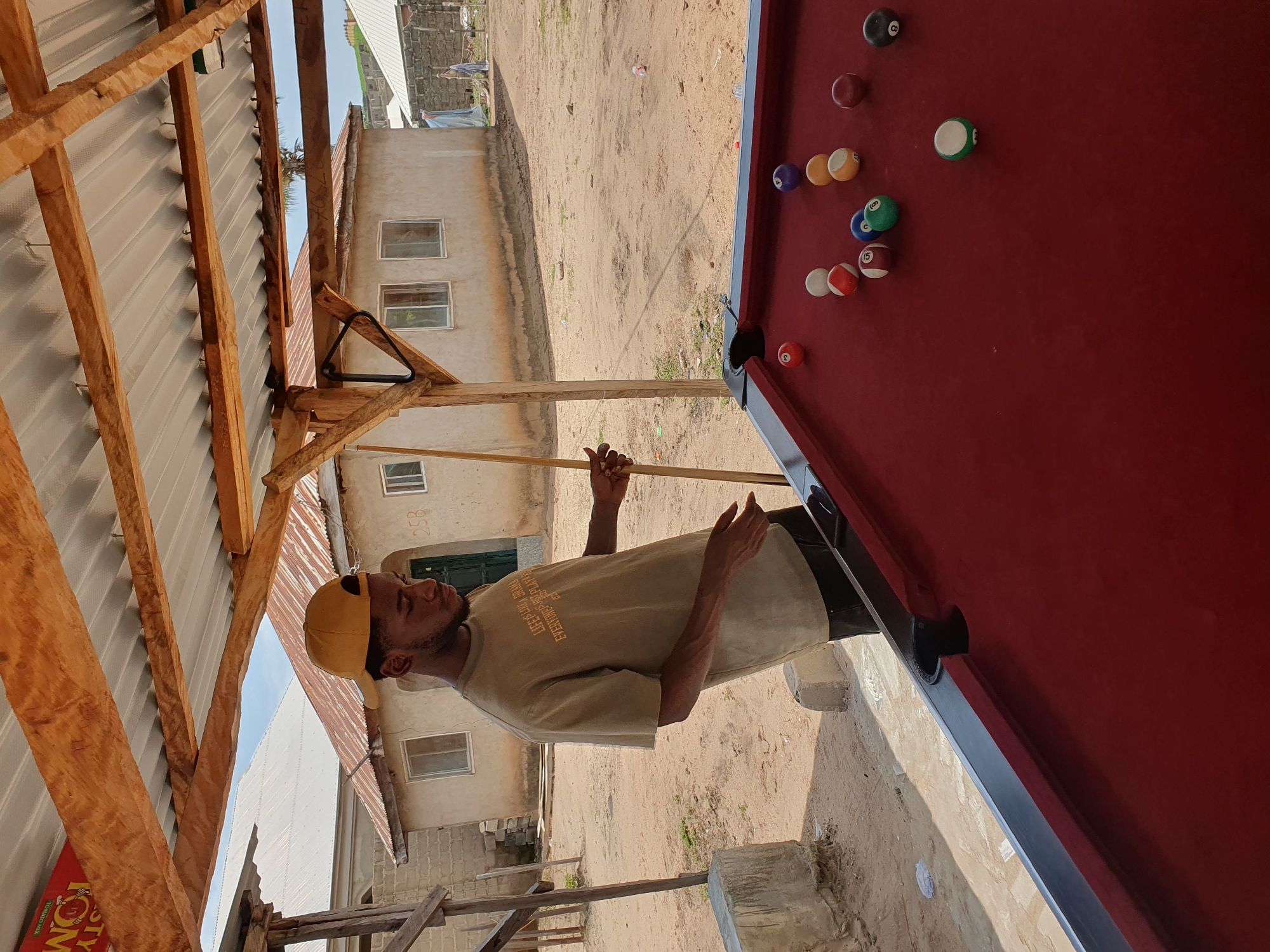 In Lokoja, Nigeria, a young man wearing a tan t-shirt and a yellow baseball cap stands outdoors under a wooden and corrugated metal roof structure. He is holding a pool cue and appears to be playing pool. A red pool table is in the foreground, with several pool balls scattered across it. The background shows a sandy ground and several buildings, suggesting a residential or commercial area. The sky is visible above the buildings. The overall scene depicts a casual outdoor setting with a game of pool in progress.