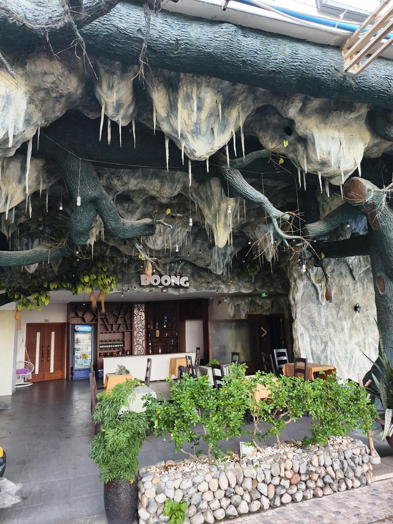 The image shows the exterior of "BOONG," a restaurant in an unspecified city. The restaurant's design is unusual, with an artificial, cave-like overhang featuring stalactite-like structures. The restaurant entrance is visible, revealing tables and chairs inside. A small stone wall with green shrubbery borders the front of the building. The setting appears to be a commercial area with a paved walkway.