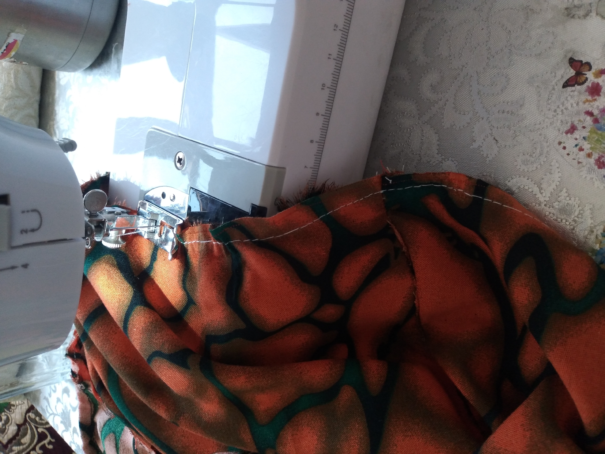 The image shows a close-up view of a sewing machine in use, with a piece of orange and green patterned fabric being sewn. The fabric is draped over the sewing machine's presser foot and needle. A white thread is visible, indicating that sewing is in progress. The sewing machine is white and appears to be a standard domestic model. The background includes a surface with a subtle embossed pattern, possibly a tablecloth, and part of a larger metallic object, likely a sewing machine accessory or another household item. There is no indication of the city of Baki, Uzbekistan, in the image.