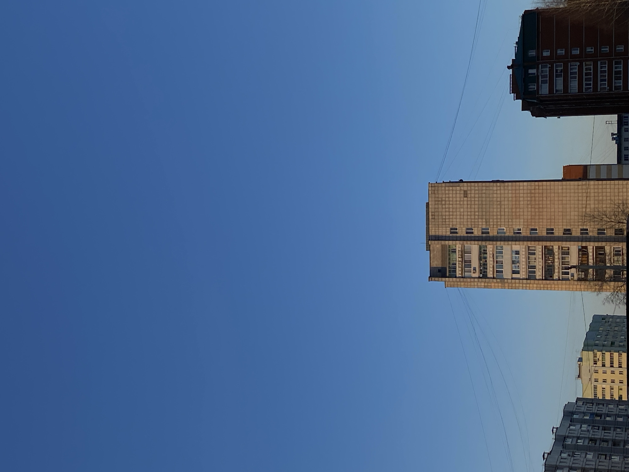 The image depicts several buildings against a clear blue sky. The buildings appear to be apartment blocks of varying heights. One of the buildings is a light-colored, multi-story structure with numerous windows. Other buildings, darker in color, are visible in the upper right and lower right corners of the image. There are also several wires stretching across the sky between the buildings. This photo was likely taken in Chaikovsky, Russia, an urban area characterized by similar architectural styles.
