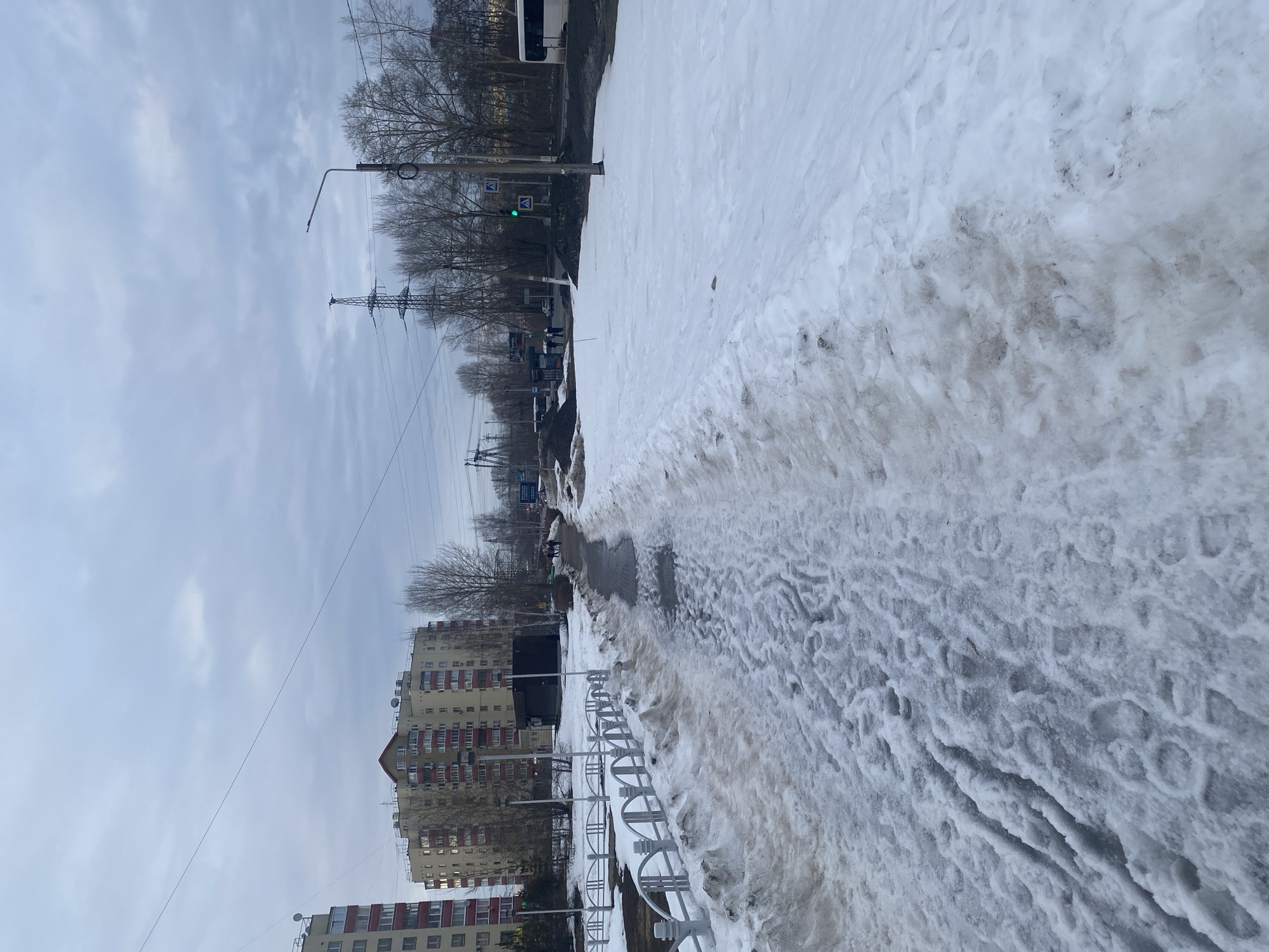 The image displays an urban environment in Chaikovsky, Russia, during what appears to be late winter or early spring, under an overcast sky. A snow-covered pedestrian sidewalk, marked by numerous footprints and areas of dirty, melting snow interspersed with wet pavement, extends from the foreground into the mid-distance on the left. This sidewalk is adjacent to a low white ornamental metal fence, beyond which lies additional dirty snow.
To the right of the pedestrian path, a broader expanse of snow covers the ground, leading towards a multi-lane road. Along this road, a bus stop shelter is visible in the mid-distance, where several indistinct individuals are present, appearing to be waiting. A white bus is partially visible on the far right. Dark piles of material, resembling gravel or dirt, are situated near the road.
In the background, two multi-story residential apartment buildings with a characteristic post-Soviet architectural style are prominent on the left. The more central building is light-colored with red window and balcony accents. Bare deciduous trees are scattered throughout the scene, indicative of the season. Utility infrastructure includes power lines spanning the frame, several utility poles, a street light on a pole, and a tall lattice-type electricity transmission tower in the center-right background. A green pedestrian crossing sign with a white 'A' symbol is visible on a pole near the road.