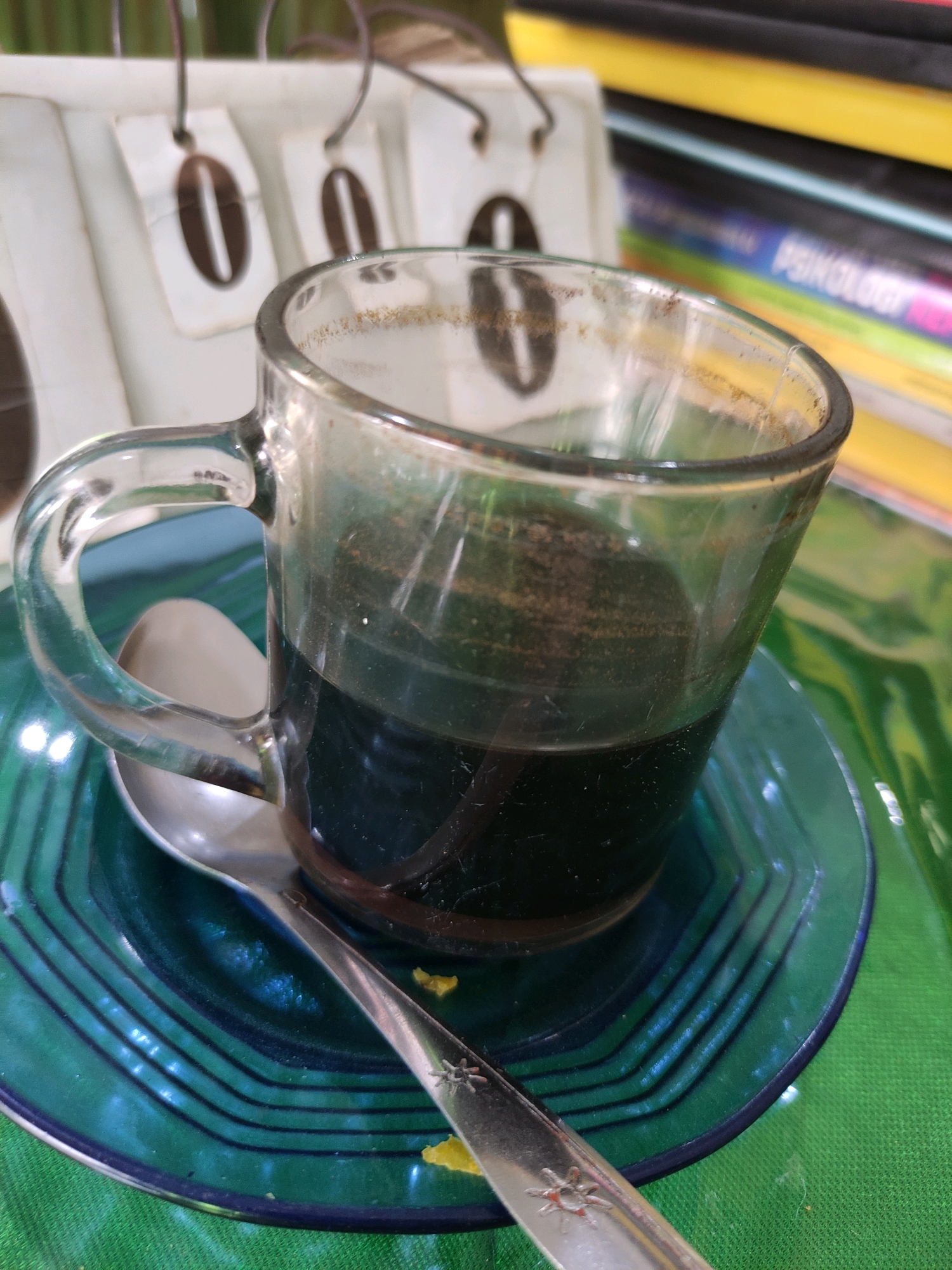 A close-up shot focuses on a half-filled clear glass mug of dark liquid, likely coffee, resting on a patterned teal glass saucer. A silver spoon is placed on the saucer, partially beneath the mug. In the background, out of focus, are several books stacked horizontally, with one visible spine showing the word "PSYCHOLOGY" in colorful lettering. To the left, a white decorative item with circular cutouts resembling coffee beans is partially visible. The overall scene is set on a green textured surface, possibly a tablecloth. The image appears to have been taken indoors, possibly in a home or a cafe setting, and is not representative of any specific location within Bulukumba, Indonesia.