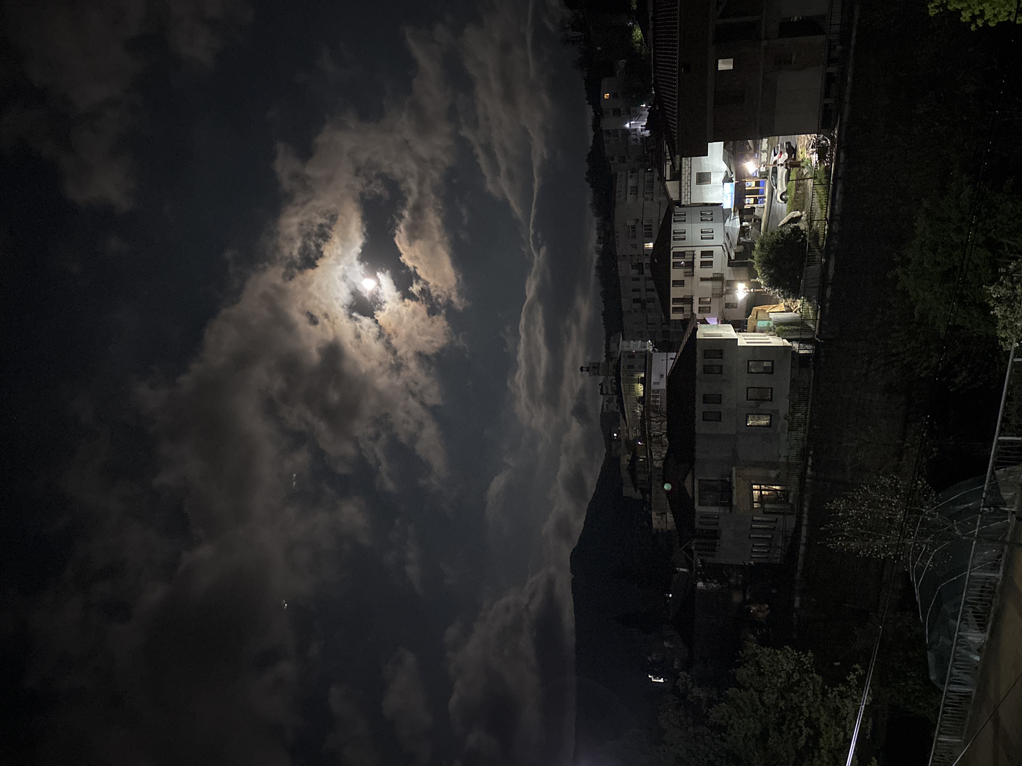 A night scene shows a partially cloudy sky with a bright moon visible through the clouds. To the right, a cluster of buildings with lit windows suggests a residential area. Vegetation is visible around the buildings. The scene is possibly Zlatograd, Bulgaria, judging by the type of buildings and landscape features. The photograph is rotated 90 degrees counter-clockwise. No people are discernible in the image.