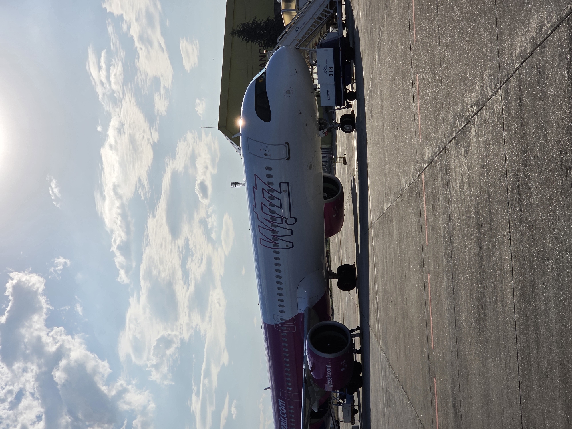 This image depicts a Wizz Air airplane parked on an tarmac at an airport. The airplane is the primary subject, prominently displayed in the center of the frame. It is a passenger jet with white fuselage and distinctive pink accents on its engines and tail. The Wizz Air logo is visible on the side of the aircraft. The scene is set outdoors, with a bright, sunny sky filled with scattered white clouds occupying the upper portion of the image. The sun is shining brightly, casting a glare on the airplane's nose and creating some lens flare. The tarmac is visible in the foreground and right side of the image, with markings indicating parking positions.
A boarding stair truck is attached to the airplane's door, suggesting that passengers may be boarding or disembarking. A few figures, presumably airport personnel or passengers, are visible near the truck and the airplane's wheels, though they are small and not clearly discernible. In the background, a building with yellow siding and a sign that appears to say "AIRPORT" is partially visible, along with some trees. The overall atmosphere appears calm and operational, typical of an airport environment. The image seems to be taken during daytime, likely late morning or afternoon, given the bright sunlight and shadows. No specific events or emotions are clearly conveyed, beyond the functional activity of an aircraft at an airport. Text visible includes "WIZZ" on the airplane and "wizzair.com" along the lower fuselage. The boarding stair truck has "313" written on a white box.