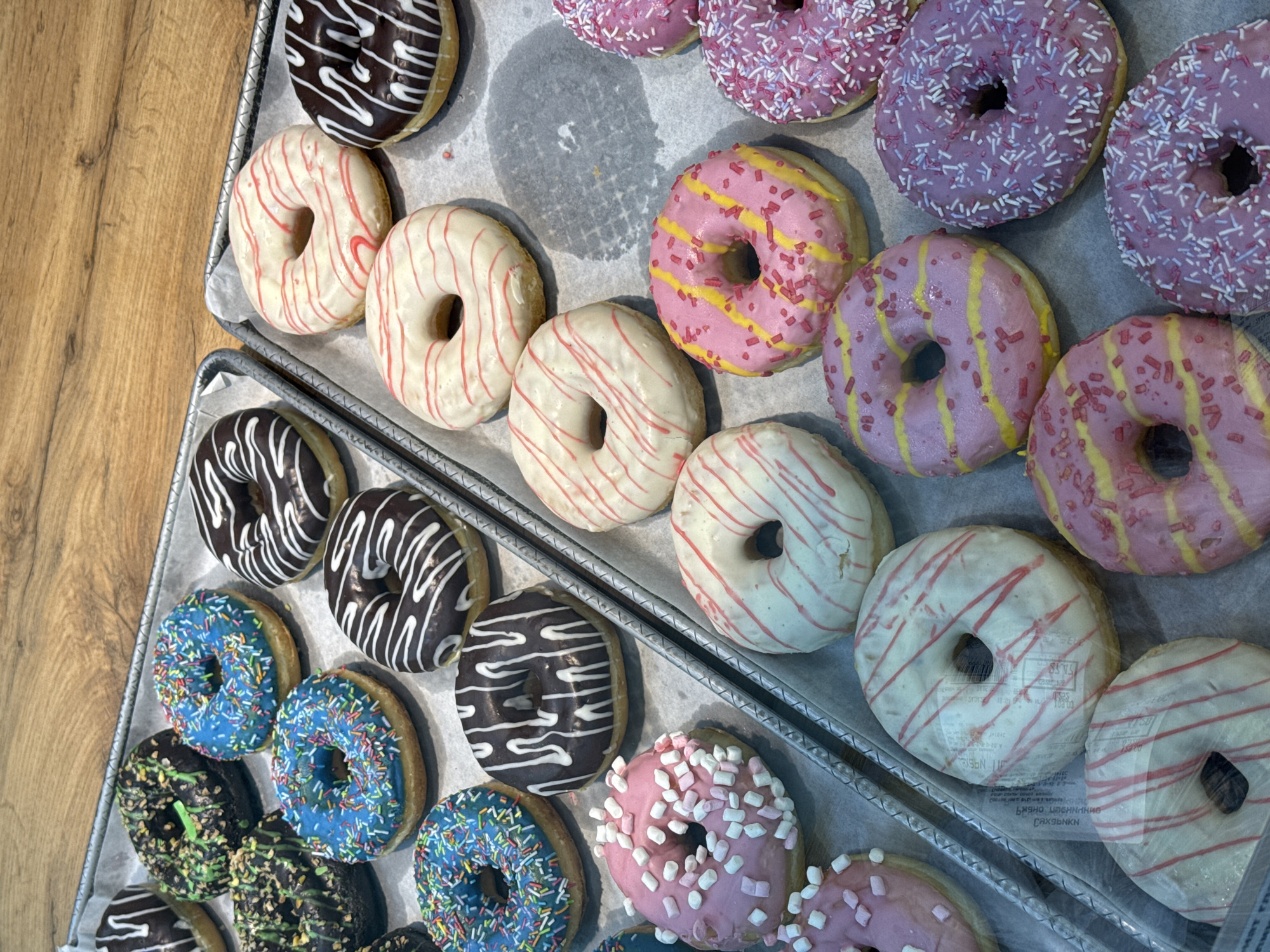 Two metal trays are filled with an assortment of donuts, arranged closely together. The trays are lined with parchment paper.
The donuts are glazed with various colors, including dark brown with white stripes, white with red stripes, blue with multi-colored sprinkles, dark green with green sprinkles, pink with white marshmallow pieces, pink with yellow stripes and pink sprinkles, and purple with white and purple sprinkles.
The trays are located in an Unknown area of Unknown.