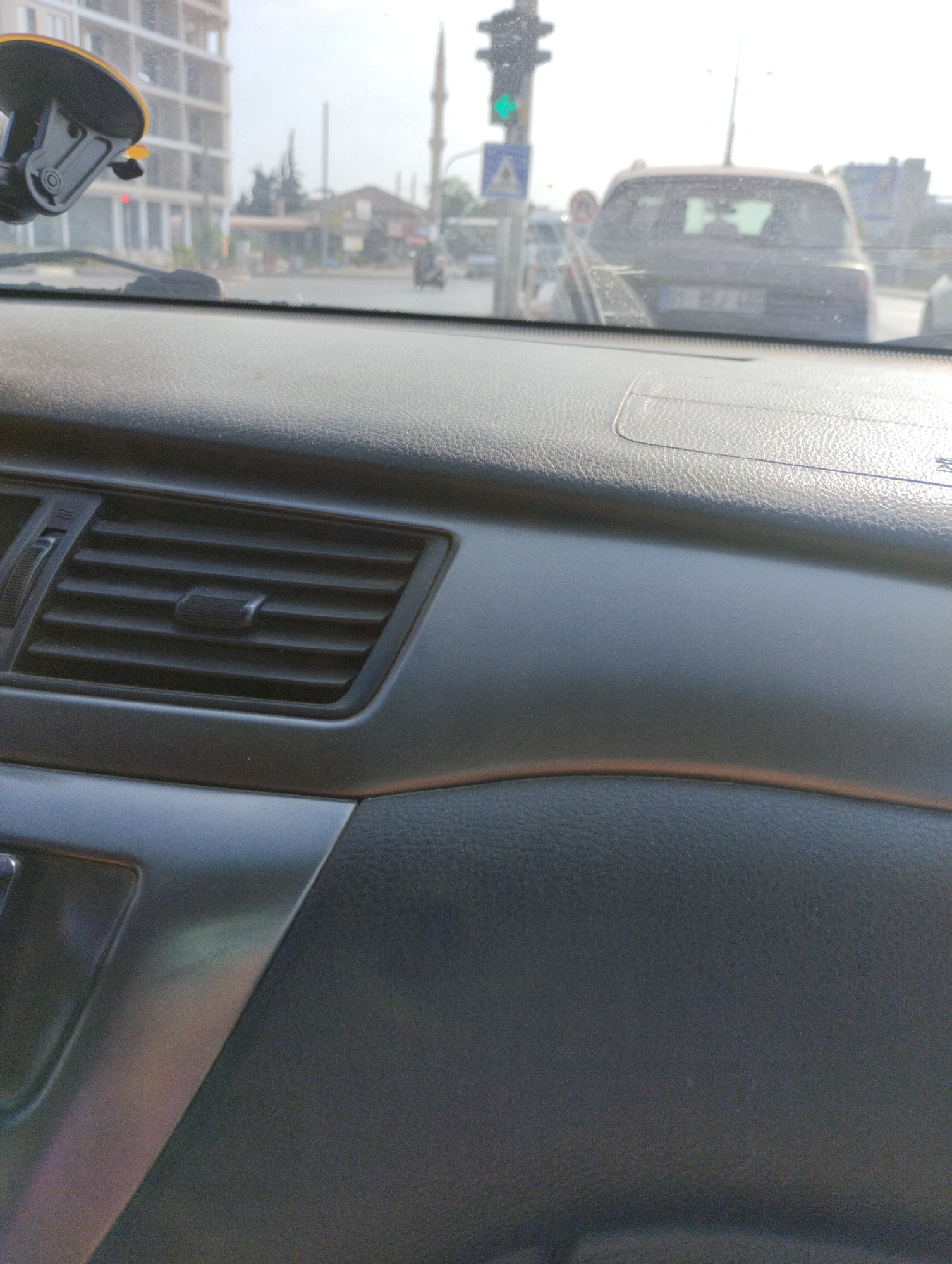 The image is a close-up, interior view from a vehicle, looking out through the windshield. The dashboard is visible in the foreground, featuring a black air vent with adjustable louvers and a textured surface. The exterior scene is slightly blurred, showing a street with traffic. A tall, modern building is visible on the left. In the center, a traffic light with a green signal is illuminated. A car with a license plate is in front of the camera's view, and further back, a mosque minaret can be seen. The overall environment suggests a sunny day in an urban setting, likely Erdemli, Turkey, given the presence of typical street infrastructure and architecture.