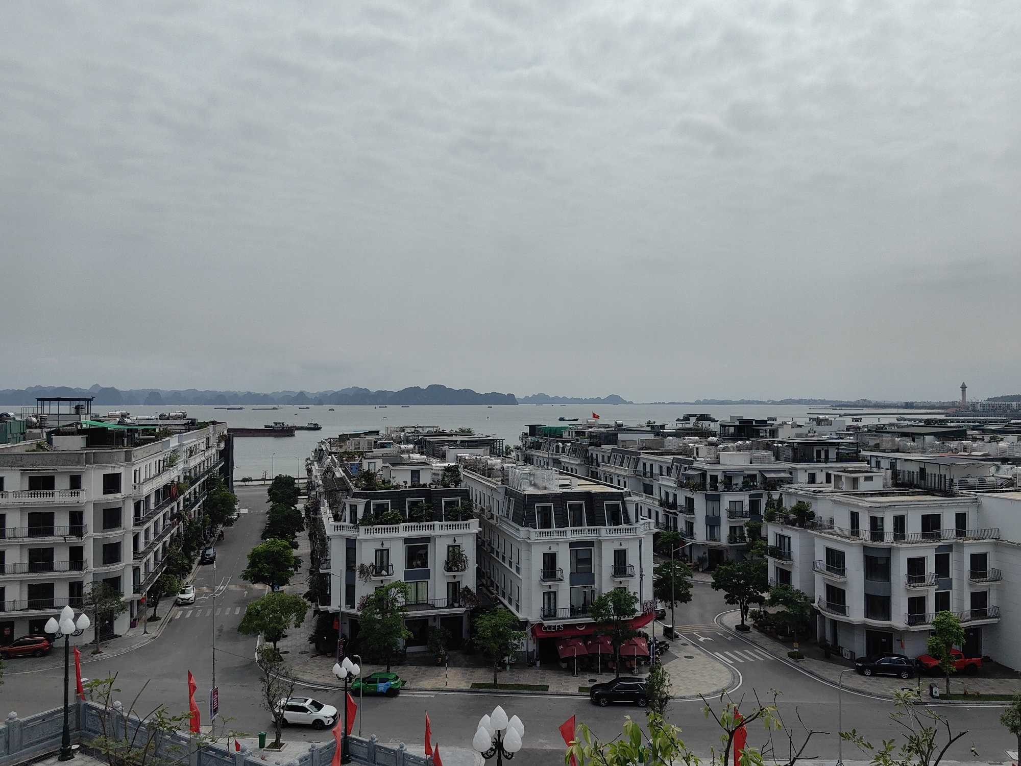 The image shows an elevated view of a district in Hong Gai Ward, Vietnam. The area is characterized by uniform, multi-story white buildings with dark roofs, many featuring balconies and decorative elements. Streets are lined with trees, and some commercial establishments are visible on the ground floors. A large body of water with several ships is visible in the background, with islands on the horizon under an overcast sky. A tall tower can be seen in the distance on the right, and red flags are planted in the foreground. Several cars are parked along the streets and intersections.