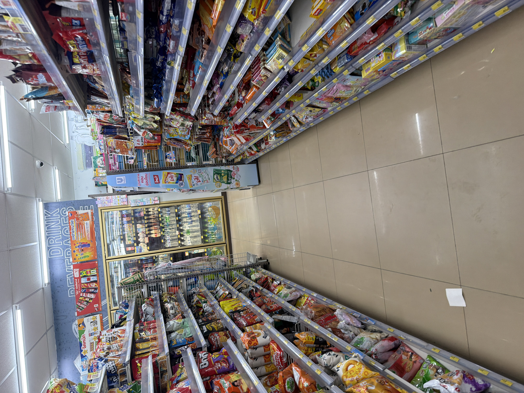 The image captures an interior view of a convenience store, likely a 7-Eleven, as indicated by the familiar shelving and product displays. The scene is set in Baan Mae Nam, Thailand. The perspective is a low-angle shot looking down an aisle, with shelves packed with various snacks and beverages lining both sides.
On the left, shelves are filled with brightly colored snack bags, including chips and crackers, with prominent Thai language branding. Some larger bags are displayed at the bottom of the shelves. Towards the back on the left, a refrigerated display unit is visible, showcasing bottled drinks. Above the refrigerated unit, a large sign with "DRINK BEVERAGES" in English and Thai script is mounted on the wall.
On the right, shelves are also laden with snacks, including boxed items and smaller bags, with a mix of English and Thai text on the packaging. The flooring is a neutral-toned tile. The lighting appears to be fluorescent overhead lights. The overall impression is a well-stocked retail environment typical of a convenience store in Thailand.
