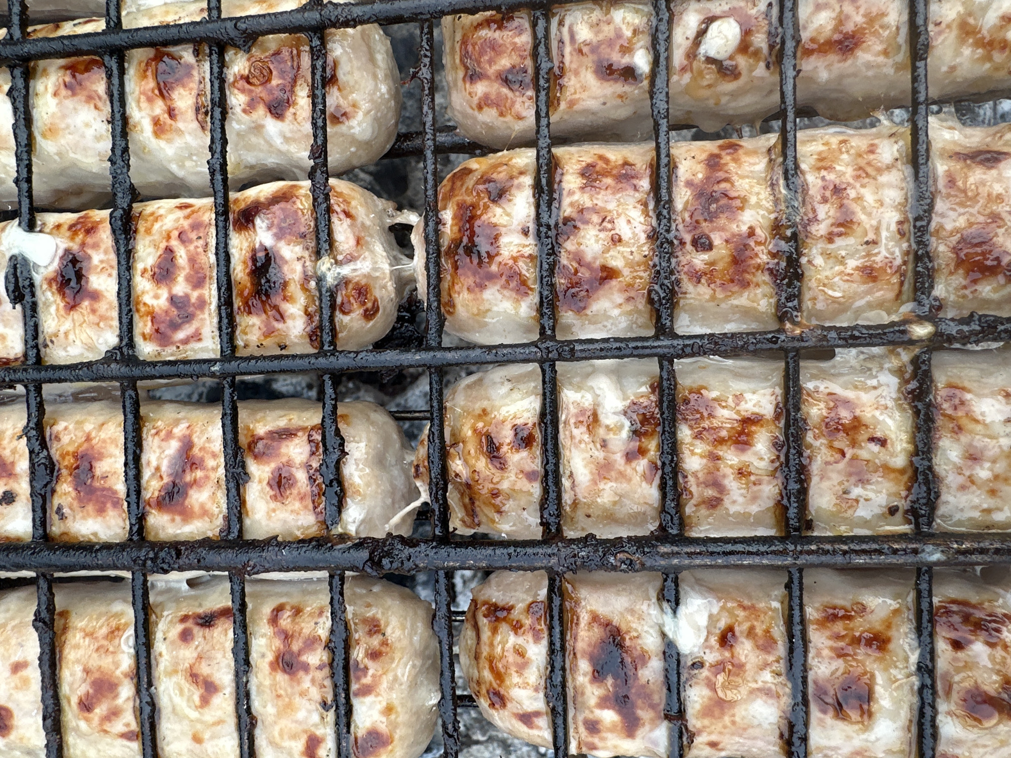 The image shows sausages grilling on a black metal barbecue grill. The sausages are pale with browned areas from the cooking process. Some sausages show signs of splitting. There are no people visible in the scene. The location is unknown.