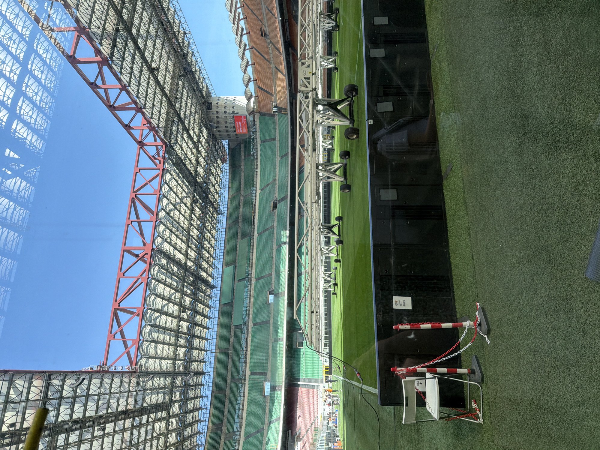 The image shows the San Siro Stadium in Milan, Italy, viewed from an elevated position, likely behind glass. The stadium features a latticework structure in the upper left, with a red support beam visible against a blue sky. The green seating tiers of the stadium run horizontally across the center of the image. There are also some ground level structures on the right side, like stage-like lights. A black monitor, and a red and white barricade with a chained chair is also visible on the right side of the frame.