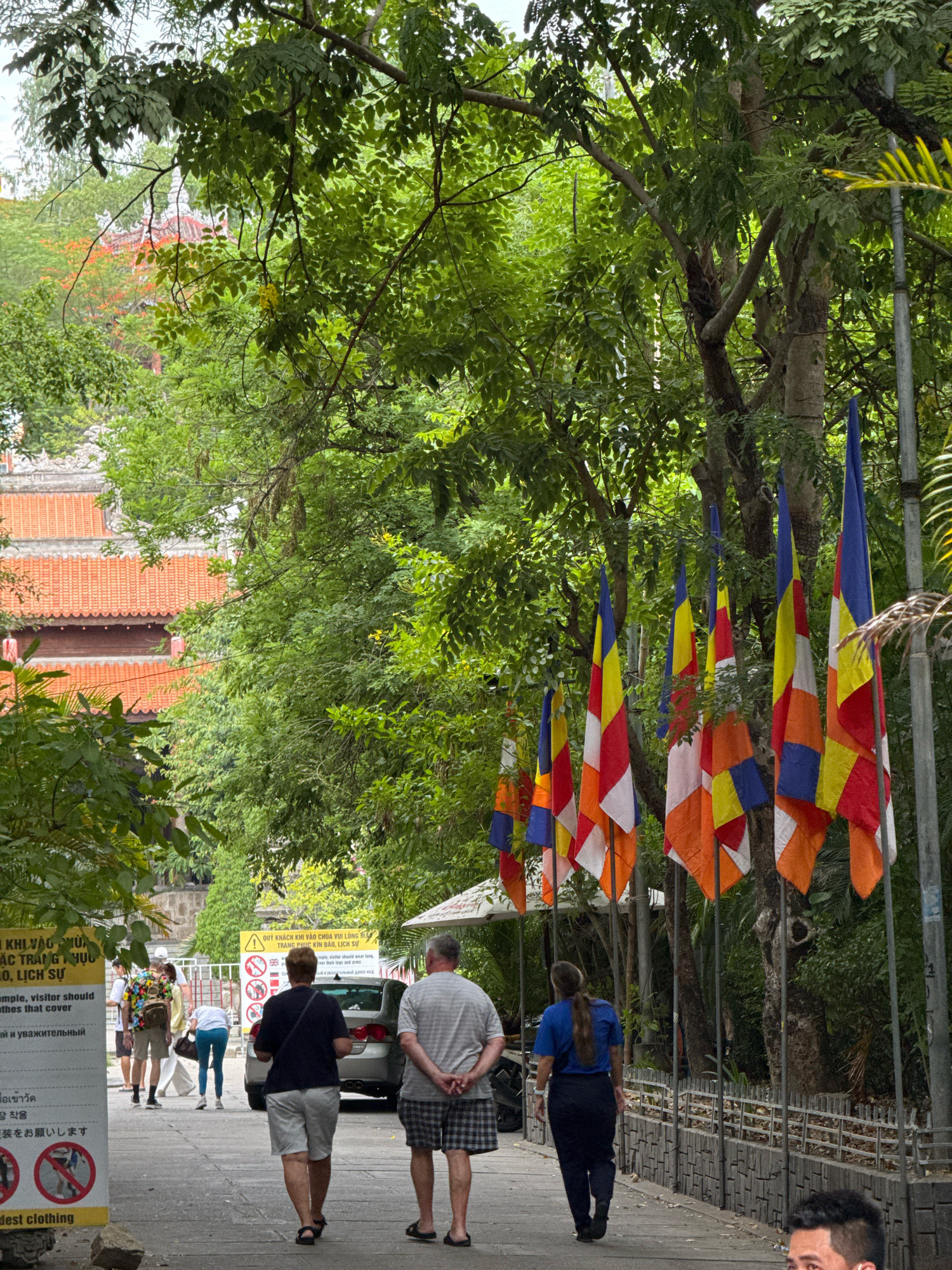 The image shows a pathway in what appears to be a temple or cultural site in Nha Trang, Vietnam. The pathway is lined with lush green trees on the left and right, with a traditional Vietnamese building featuring an orange tiled roof visible through the foliage in the background. A row of Buddhist prayer flags, with their distinct color combinations of blue, yellow, red, white, and orange, are prominently displayed on the right side of the pathway. Several people are walking along the pathway. In the foreground, a couple in casual attire walks away from the camera, followed by a woman in a blue shirt and dark pants. To the left, a group of people, including a person with a colorful backpack, are walking towards the camera, with a sign in Vietnamese and English visible, indicating a dress code that requires visitors to cover their shoulders and knees. The overall atmosphere is one of a peaceful, cultural landmark with visitors observing the rules and customs.