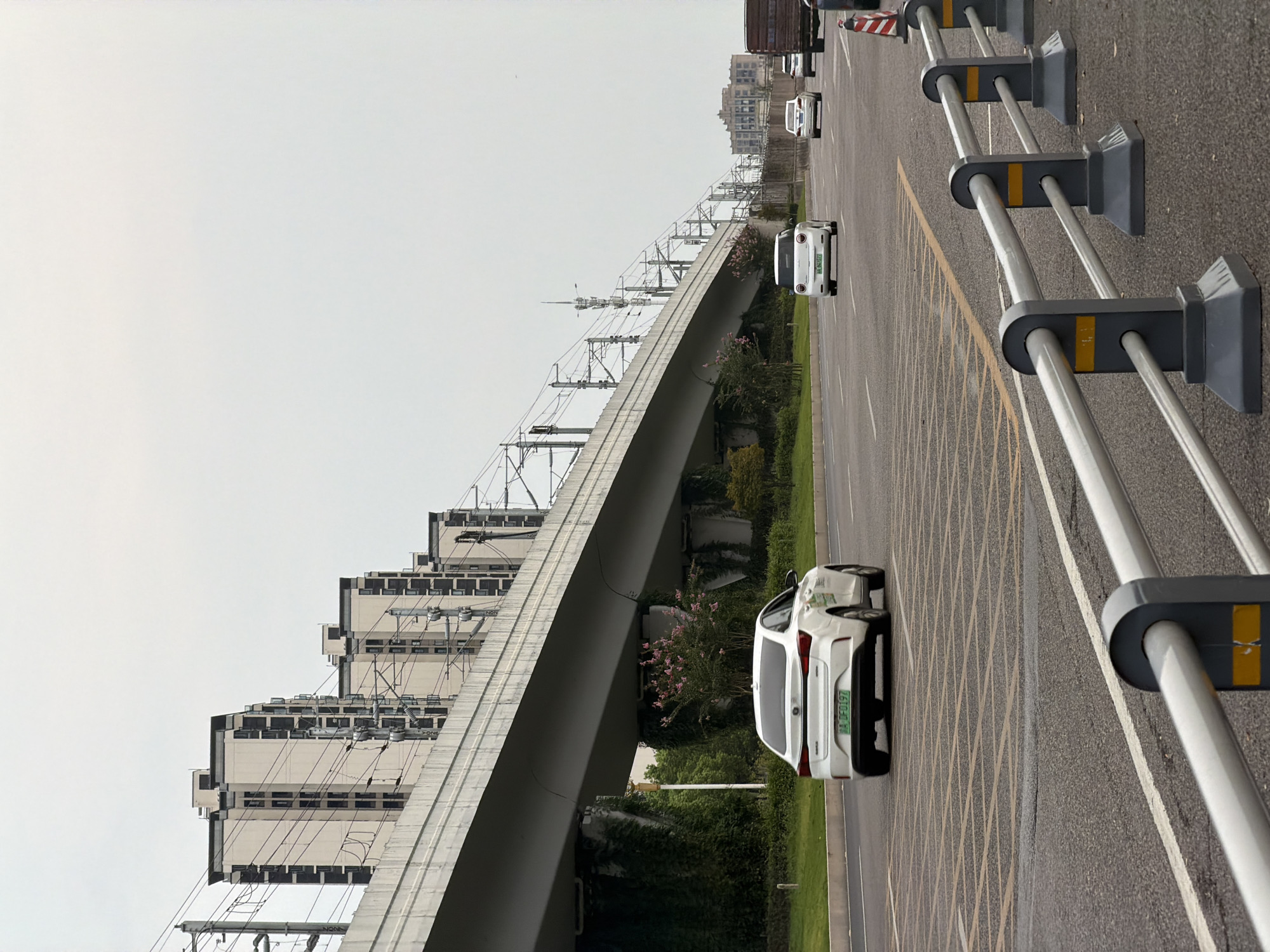 This image captures a view of a road in the city of Jiangjin, China, beneath an elevated structure that appears to be part of a railway or an overpass. Several modern, multi-story buildings are visible in the background, lined up along the elevated structure. The sky is overcast, contributing to a soft, diffused light.
On the road, four vehicles are present. In the foreground, a white sedan with a green license plate is driving away from the camera. Further down the road, two other white sedans are also in motion. On the right side of the road, a large truck with a brown cargo bed is partially visible.
The road is paved with asphalt and features multiple lanes. Yellow and white markings are painted on the road surface, indicating lanes and traffic flow. A section of the road in the foreground has a grid pattern of yellow lines, possibly marking a special zone or intersection.
Along the right edge of the road, a metal barrier system with gray posts and silver horizontal rails is installed. The posts have dark gray bases and yellow accent markings. Orange and white traffic cones are also visible near the barrier, suggesting potential road work or traffic management.
The elevated structure is supported by large, vine-covered concrete pillars. Greenery, including bushes and trees with pink blossoms, grows alongside the road and under the overpass. Power lines and associated infrastructure are mounted on poles alongside the elevated structure, indicating an electrified transportation system. The scene depicts a typical urban environment with infrastructure and transportation elements.
