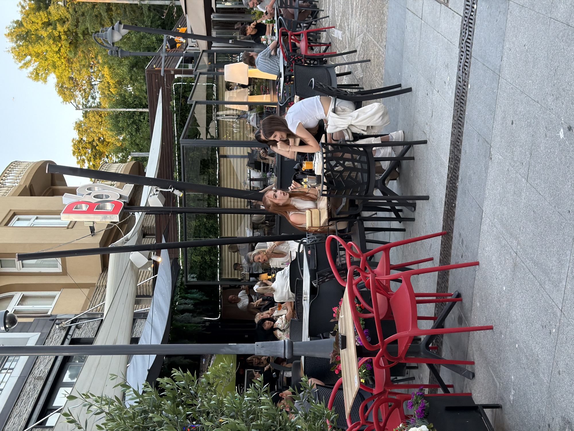 The image shows an outdoor seating area of a restaurant in Burgas, Bulgaria. Several people are seated at tables, some are talking, eating, or drinking. In the foreground, a table with red chairs is visible. In the background, the facade of a building with a balcony is seen, along with trees and streetlights. The atmosphere appears to be relaxed, with people enjoying the evening outdoors.