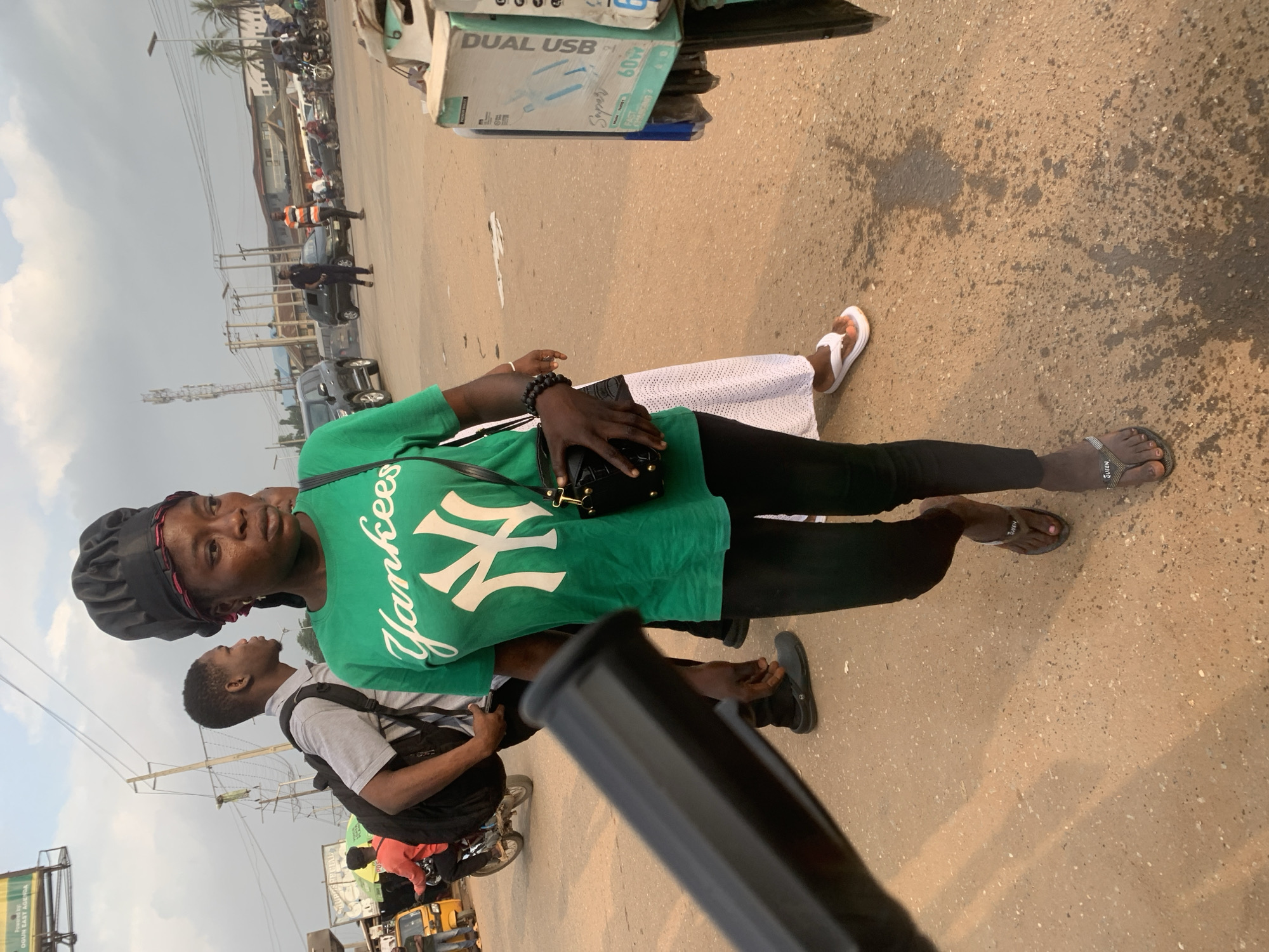 The image captures a street scene in Shagamu, Nigeria, on a cloudy day. In the foreground, a Black woman is walking towards the right of the frame. She is wearing a green t-shirt with a white "Yankees" logo and a black skullcap. She has a black handbag with gold hardware slung across her body and is wearing black leggings and flip-flops. To her left and slightly behind her, a young Black man with a backpack walks in the same direction. In the background, several vehicles, including cars, a tricycle, and motorcycles, are present, suggesting a busy urban environment. The ground is paved with asphalt and shows signs of wear and tear, with some water stains visible. The overall impression is of a typical street scene in a Nigerian city.