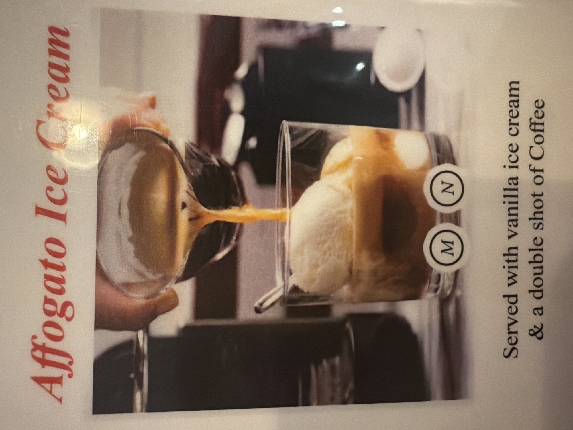 The image shows a close-up of an Affogato dessert being prepared. A hand pours coffee from a small clear glass pitcher into a taller clear glass containing vanilla ice cream. The dessert is identified as "Affogato Ice Cream" in red text on the left side of the image. Text on the right states "Served with vanilla ice cream & a double shot of Coffee". The location appears to be London, United Kingdom, but there is no other information about the location.