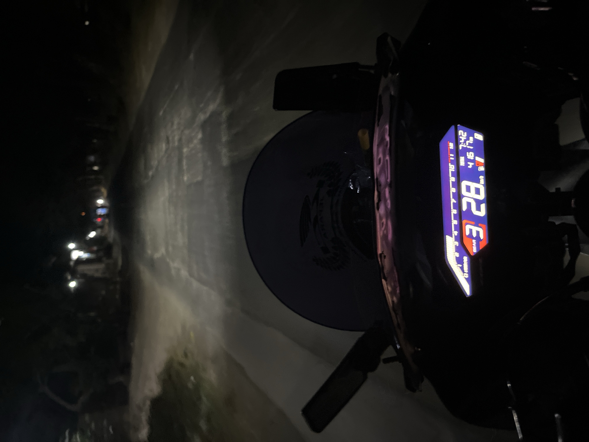 The image depicts a nighttime view from the perspective of someone riding a motorcycle on a road, likely in or near Pecangaan, Indonesia. The road ahead is dimly lit, with the motorcycle's headlights illuminating a stretch of asphalt. On the right side of the road, there are indications of vegetation and possibly some small structures or lights in the distance. The foreground shows the motorcycle's dashboard, which includes a digital display showing the speed as "28 km/h", the current gear as "3", and the time as "7:42". The overall impression is one of a quiet, possibly rural, nighttime journey.