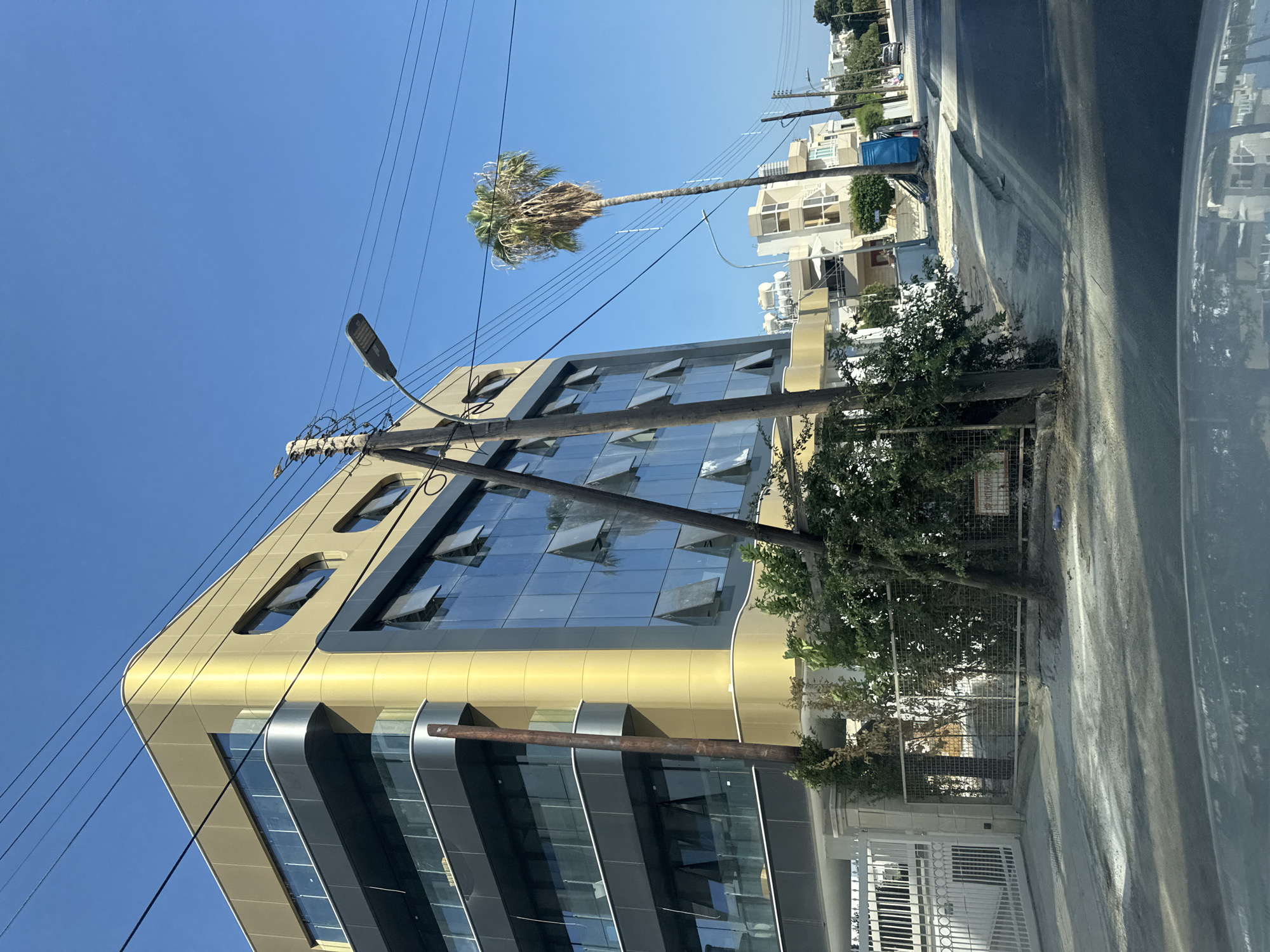 The image shows a street scene in Limassol, Cyprus. A modern, multi-story building with a gold and dark grey facade dominates the left side of the frame. The building features large glass windows, some of which are open. Several utility poles with overhead wires and a street lamp are situated in front of the building. To the right, a tall palm tree stands next to a street. Residential buildings and more trees are visible in the background. The road in the foreground appears to be asphalt, with some wear and tear. The sky is clear and blue. There are no people visible in the image. The scene suggests an urban environment on a sunny day.