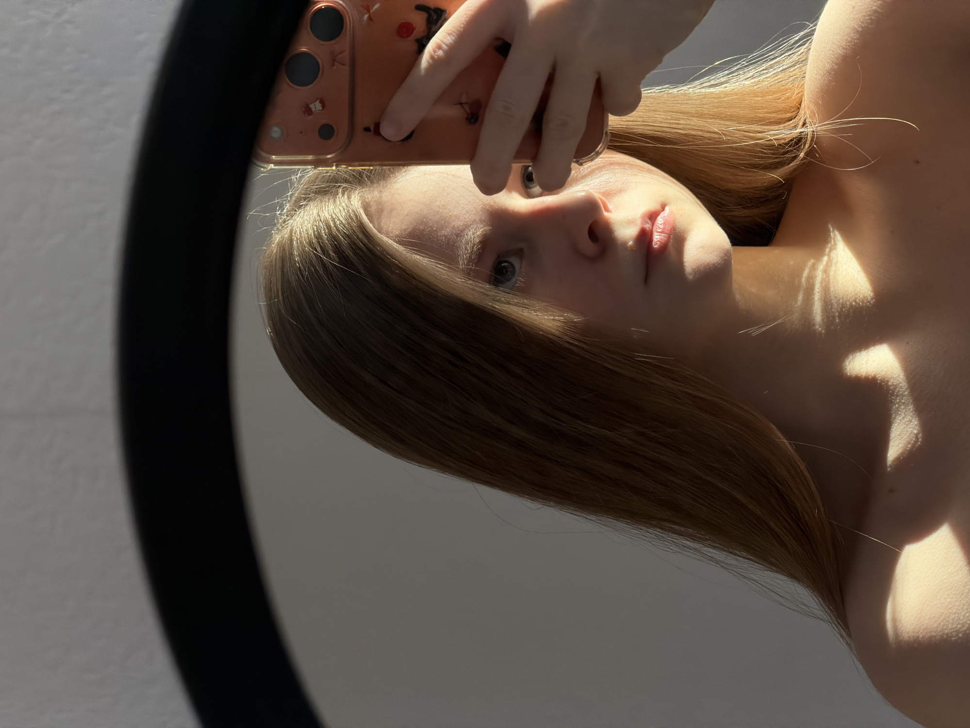 This media file is a mirror selfie of a young woman, presented in a portrait orientation but originally captured with a significant tilt. The woman, with long, straight, blonde or light brown hair, is looking directly into the mirror with a neutral expression. Her face and hair are brightly illuminated by strong, directional sunlight, creating highlights and soft shadows, and a warm glow on her hair. Shadows from what appear to be window frames or blinds are cast across her bare shoulder and upper chest.
She is holding a smartphone with a light orange or rose gold case, which has multiple camera lenses visible. The phone case is adorned with several small, dark stylized decals, including a faint letter "K" and a small animal figure (possibly a cat or bear) near the camera module. Her right hand, holding the phone, partially obscures the upper left portion of her face in the reflection. The scene's background in the reflection is a plain, light-colored wall, and a thick, curved black frame on the left side confirms it is a mirror selfie. The strong natural light indicates a daytime setting. No specific location cues are visible beyond an indoor environment.