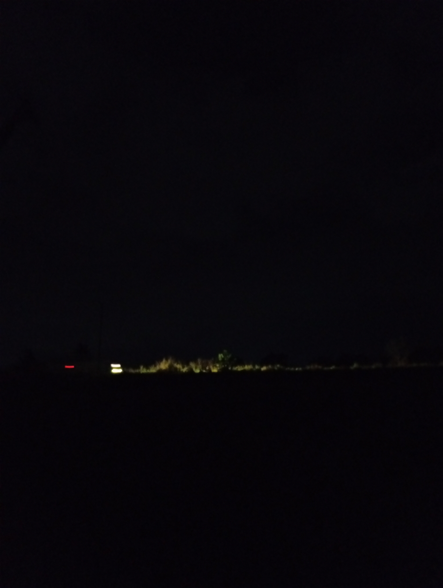 The image is a dark, horizontal shot of a landscape at night. In the distance, a faint line of lights suggests a road or a settlement. Two sets of lights are visible: a horizontal red light on the left, and two parallel yellow lights on the right, possibly indicating vehicles. The ground is dark and indistinct, with some textured vegetation visible along the horizon, illuminated by the faint lights. The sky is entirely black. The image does not provide any specific details about the city or its location.