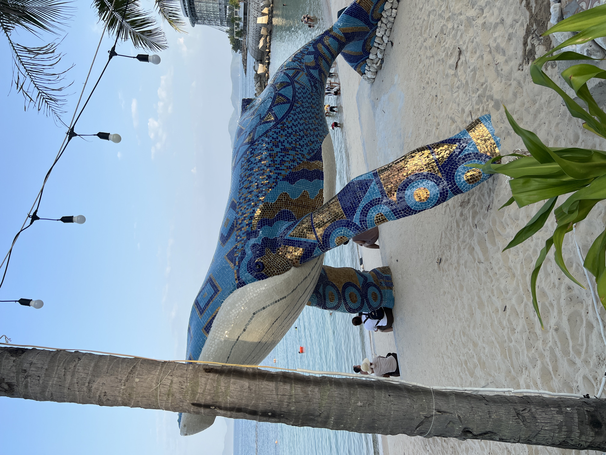 A mosaic sculpture of a whale is displayed on a sandy beach, likely in Nha Trang, Vietnam, given the architectural style of the building in the background and the tropical foliage. The whale sculpture, predominantly blue with gold accents in intricate patterns, is positioned on the sand with its body elevated by two legs, extending towards the ocean.
The ocean is visible in the background, with calm, turquoise water and a clear blue sky overhead. Several people are present in the scene. In the immediate foreground, near the left side of the image, a palm tree trunk is prominently featured, with white electrical cables running along its side and connecting to a string of lights above.
Further back on the beach, several individuals are seated or standing. Some are facing the ocean, while others are interacting with the whale sculpture. In the water, a few people are wading or swimming. The environment suggests a public beach or resort area, with some rocky formations and a large, modern building visible in the distance, which appears to be a hotel or resort complex. The overall scene depicts a leisure setting with art and natural beauty coexisting.