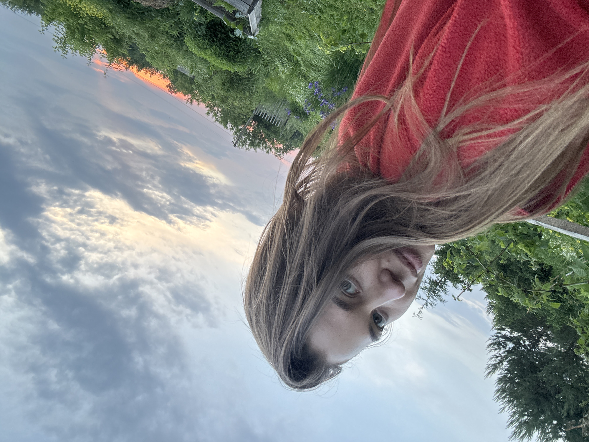 A woman with long, light brown hair is taking a selfie outdoors. She is wearing a red, fuzzy hooded garment. Her face is partially visible on the left side of the frame, with her head tilted back and to the right. She is looking towards the camera with her eyes open. The background consists of a lush, green landscape with trees and foliage. In the distance, there is a wooden fence and a glimpse of a pale blue sky with scattered clouds, tinged with the orange and pink hues of a sunset. The scene suggests an outdoor location, possibly a garden or rural area, with the natural light indicating either early morning or late evening. The image appears to be taken in Сульмаш, Russia, given the context.