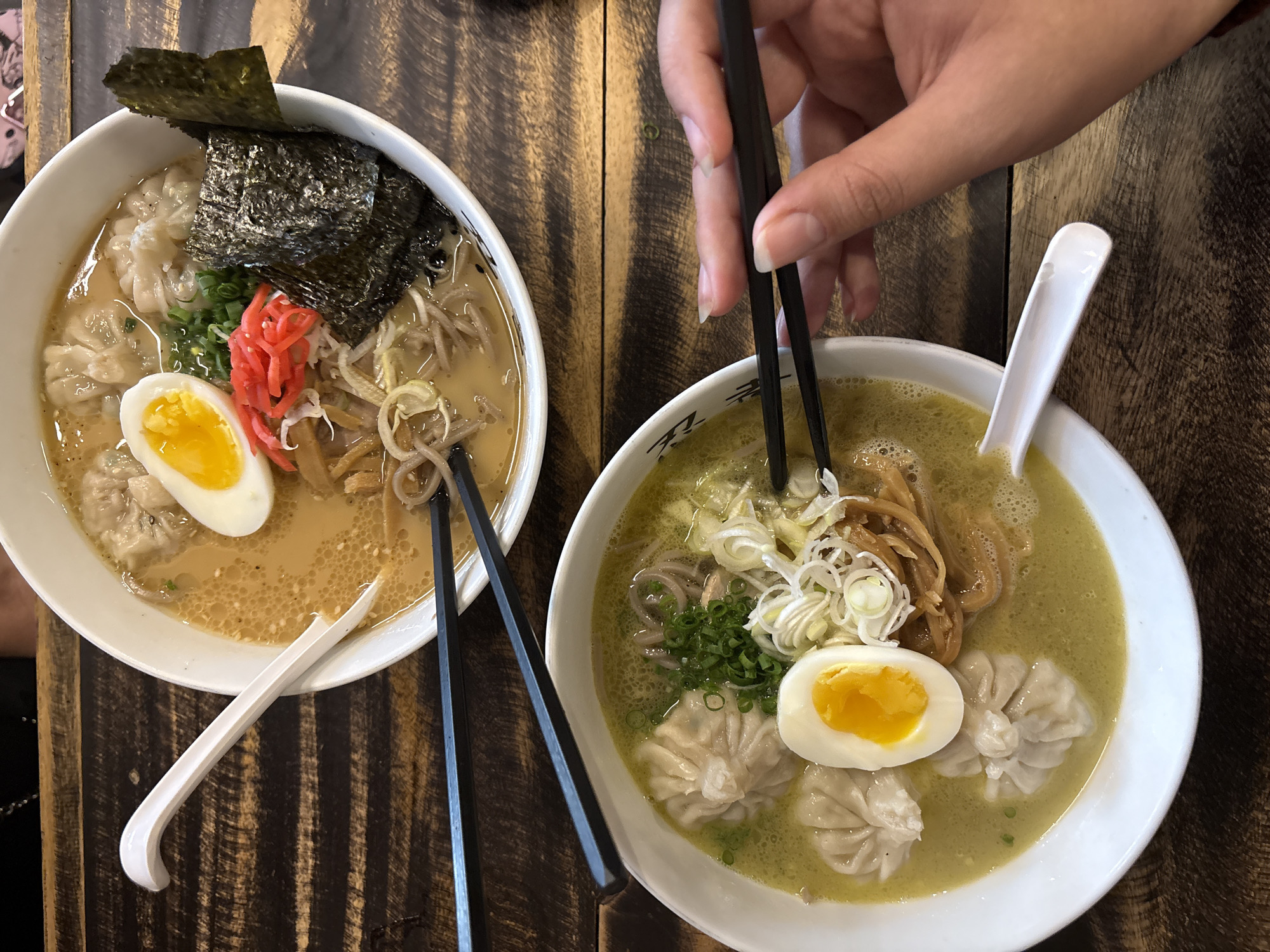 Two bowls of ramen are presented on a wooden table. The ramen in the upper bowl is a lighter, creamy broth with wontons, half of a hard-boiled egg, bamboo shoots, seaweed, and red pickled ginger. The lower bowl contains a greenish broth, also with wontons, half of a hard-boiled egg, green onions, and bamboo shoots. Black chopsticks are positioned over both bowls, and a hand is seen holding chopsticks over the lower bowl, about to pick up some noodles. A white spoon rests in the lower bowl. The background suggests an indoor setting. The image does not contain any specific visual cues to determine its exact location within Chiang Mai City Municipality, Thailand.