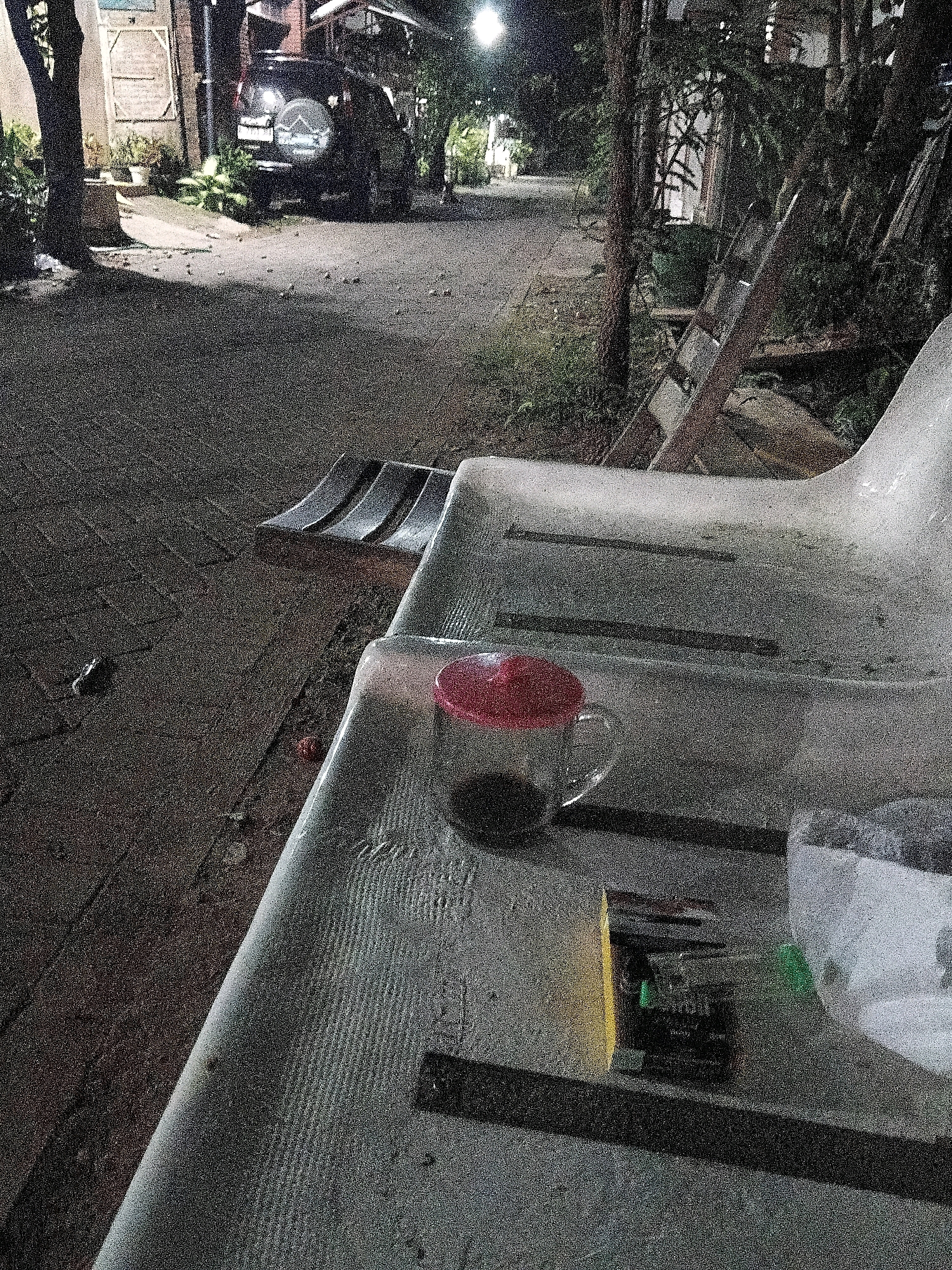 A low-angle, grainy nighttime photo captures a public street and a bench in Cikal, Indonesia. A dark-colored SUV is parked on the street, its rear visible. The bench, made of a light-colored plastic material with ribbed detailing, occupies the foreground. On the bench sits a clear glass mug with a red lid, containing a dark liquid, likely coffee or tea. Next to the mug is a pack of cigarettes and a small, white, bag-like object. A ladder leans against some foliage in the background. The scene is illuminated by ambient street lighting, casting long shadows and giving the image a muted, nocturnal atmosphere.