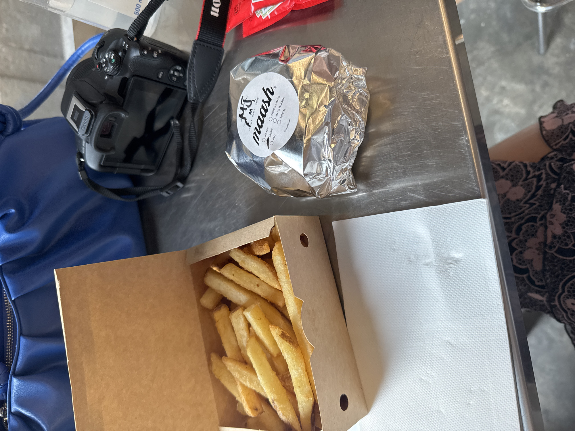 The scene is a photograph taken at a table in what appears to be a restaurant. The table has a stainless steel top, on which several objects are placed. There is a black Canon DSLR camera with its strap, a burger wrapped in silver foil with a sticker that says "maash" and flavor options, a packet of condiments, a cardboard container filled with french fries, and a stack of white napkins. Part of a blue purse is visible to the left. The presence of "maash" suggests that the location may be in Barcelona, Spain, given the branding on the burger wrapping. A person is partially visible on the right, with a patterned dark-colored sleeve. The setting appears to be indoors due to the controlled lighting and table surface.