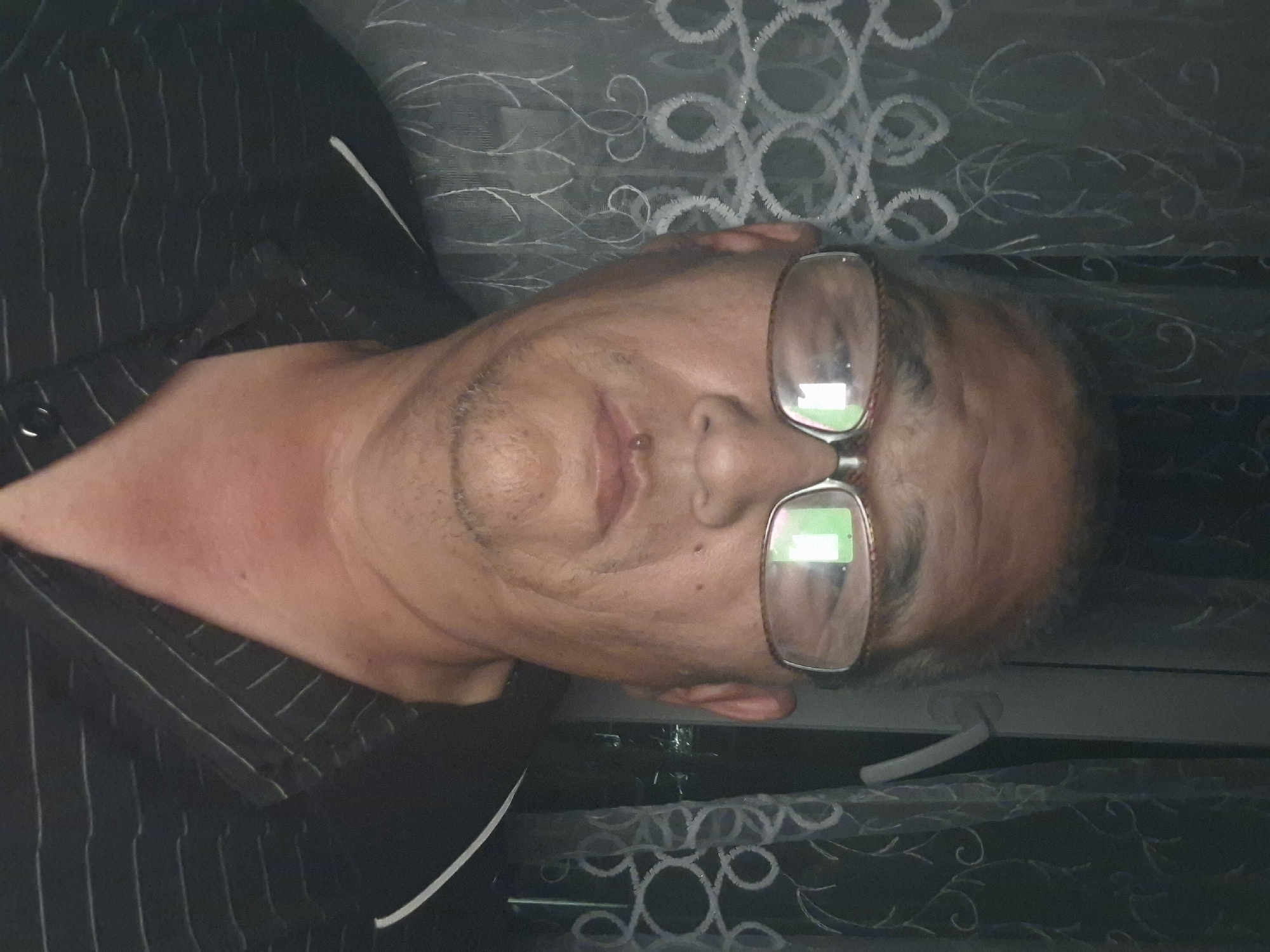 An elderly East Asian male, wearing glasses and a dark, pinstriped collared shirt, is captured in a close-up portrait. He has a small piercing on his lower lip and is looking directly at the camera. The background consists of sheer, patterned curtains, suggesting an indoor setting, possibly a residence. The lighting is dim, with the curtains allowing some light to filter through from behind, creating a softly lit, somewhat diffused atmosphere. The image does not contain any visual cues that would allow for the identification of its location in Almaty, Kazakhstan.