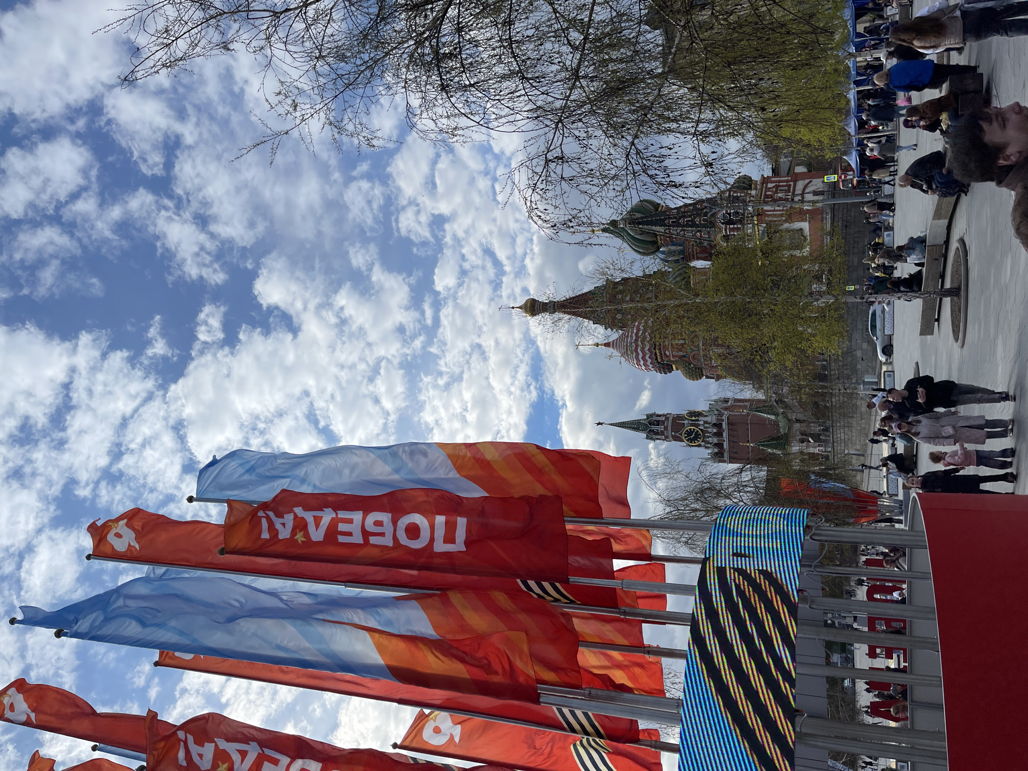 The image depicts a sunny day in Moscow, Russia. The focus is on several flags, predominantly red and blue, with a white inscription. In the background, the distinctive architecture of the Red Square is visible, including the towers of Saint Basil's Cathedral and the Spasskaya Tower of the Kremlin. People are visible walking on a path in the background. Bare branches of trees are visible above and around the towers. The sky is blue with fluffy white clouds. The image is oriented with the flags at the bottom and the sky at the top.