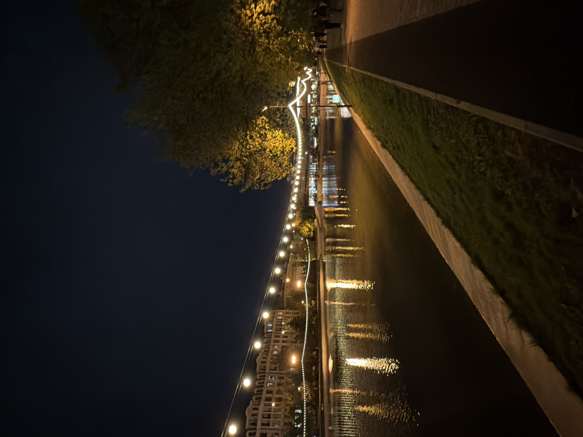 At night, a waterway runs through Kazan, Russia. Strings of lights hang along the side of the waterway. Buildings stand on the opposite bank of the waterway, also illuminated by lights. The lights reflect in the water. On the right of the waterway, there is a path with grass growing. A few people are walking on the path in the distance. The sky above is dark. A tree is on the left side.