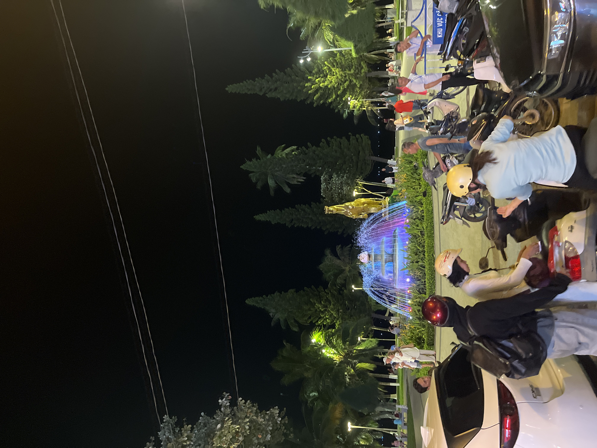The image depicts an evening scene in Nha Trang, Vietnam, likely at a public gathering space. In the foreground, several people are interacting with motorcycles. A person wearing a red helmet and a backpack is standing next to a white car, while another person in a yellow helmet is seated on a motorcycle. Further back, a colorful illuminated fountain with a golden horse statue is visible amidst lush greenery and palm trees. Numerous individuals are seen walking and socializing in the background, with streetlights providing illumination. The overall atmosphere suggests a lively, communal area, possibly a promenade or park, during nighttime.