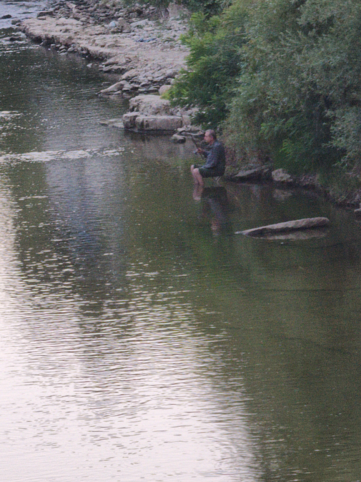 A man is sitting on the bank of a river, fishing. His legs are submerged in the water. He is wearing a dark-colored shirt and shorts. The river is calm with reflections of the trees on the far bank. The riverbank is rocky with some vegetation. The scene appears to be in a natural setting, possibly on the outskirts of Lovech, Bulgaria, near the Osam River.