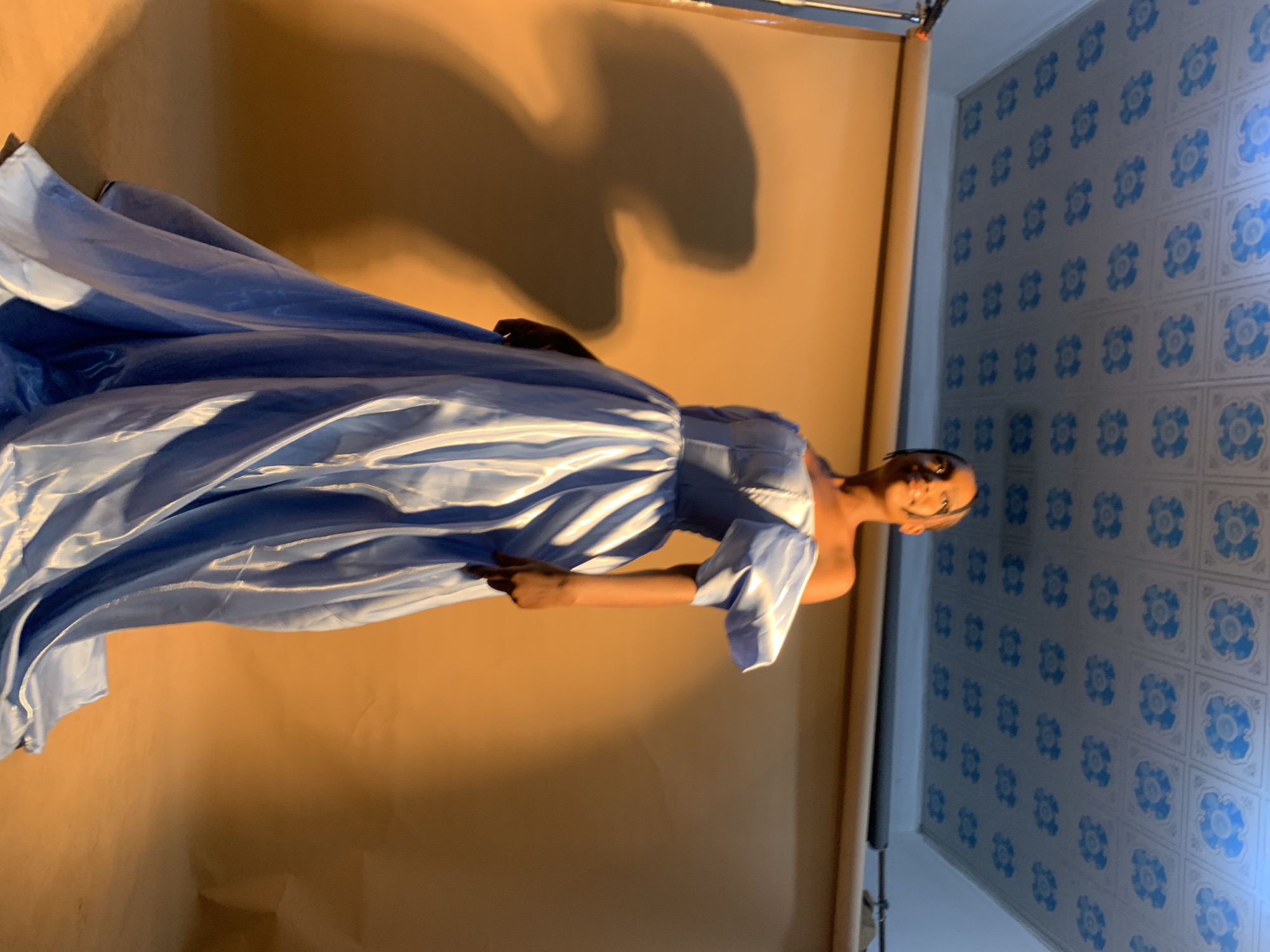 A young woman with dark hair pulled back stands on a beige backdrop in a studio. She is wearing a voluminous, off-the-shoulder light blue gown with puffy sleeves and a full skirt that trails on the floor. The fabric of the dress has a subtle sheen. Her head is tilted slightly upwards, and she is looking to her left. A shadow of a person is cast on the backdrop to her left. The ceiling above is tiled with a repeating pattern of blue floral motifs on a white background. The scene appears to be lit by warm, directional lighting, creating a strong contrast between light and shadow. There is no indication of the location being Gura Top, Nigeria.
