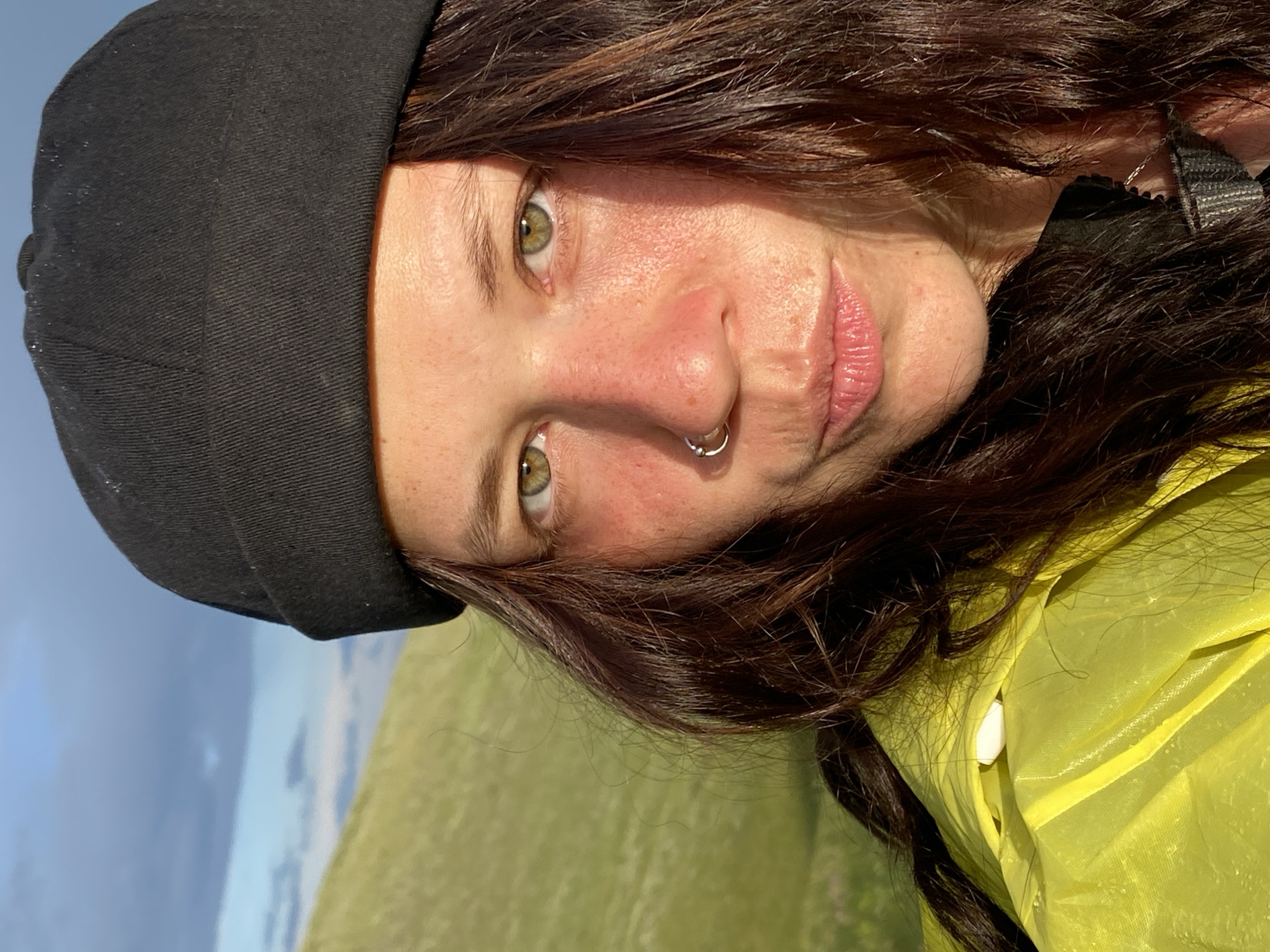 An extreme close-up selfie captures a person's face and upper body against a blurred background of a grassy hillside and sky. The person, wearing a black beanie and a bright yellow rain jacket, is looking directly at the camera. Their face is illuminated by warm, golden light, highlighting their green eyes, freckles, and a silver hoop nose ring. Their lips are slightly parted, and their dark, wavy hair frames their face. The background suggests an outdoor, possibly mountainous or hilly, environment, consistent with the location of Верхнеозёрное, Russia. The overall impression is of a casual outdoor portrait taken in natural light.