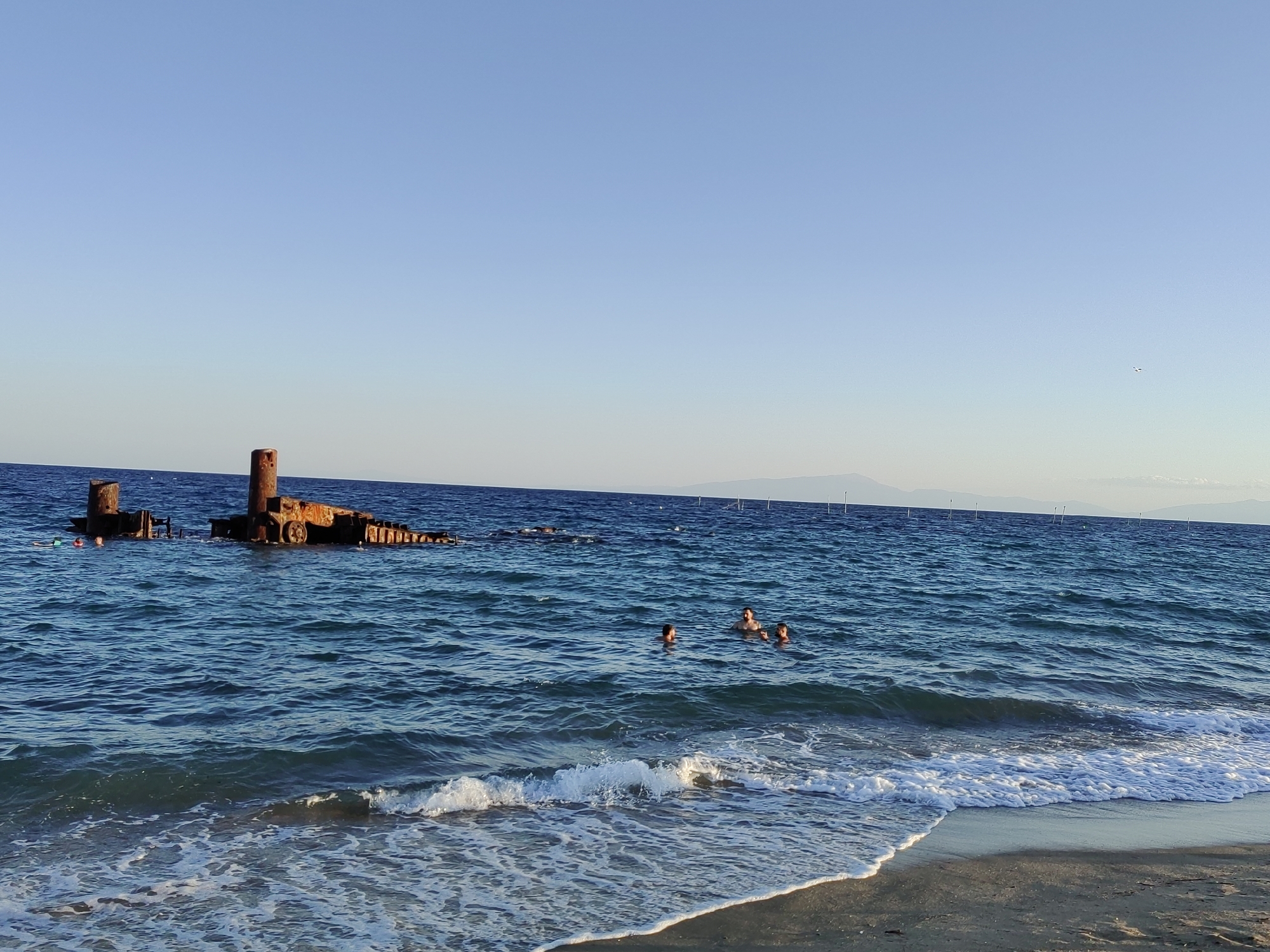 A serene beach scene in Perea, Greece, unfolds under a clear blue sky. The turquoise waters of the Aegean Sea gently lap onto a sandy shore, creating white foam. In the distance, a rusty shipwreck, partially submerged, juts out of the water, serving as a stark contrast to the tranquility. Several individuals are visible in the sea, their heads bobbing above the surface as they enjoy a swim. The coastline stretches out, with mountains forming a hazy backdrop against the horizon. The overall impression is one of a peaceful summer day at the seaside, punctuated by the presence of the historical shipwreck.