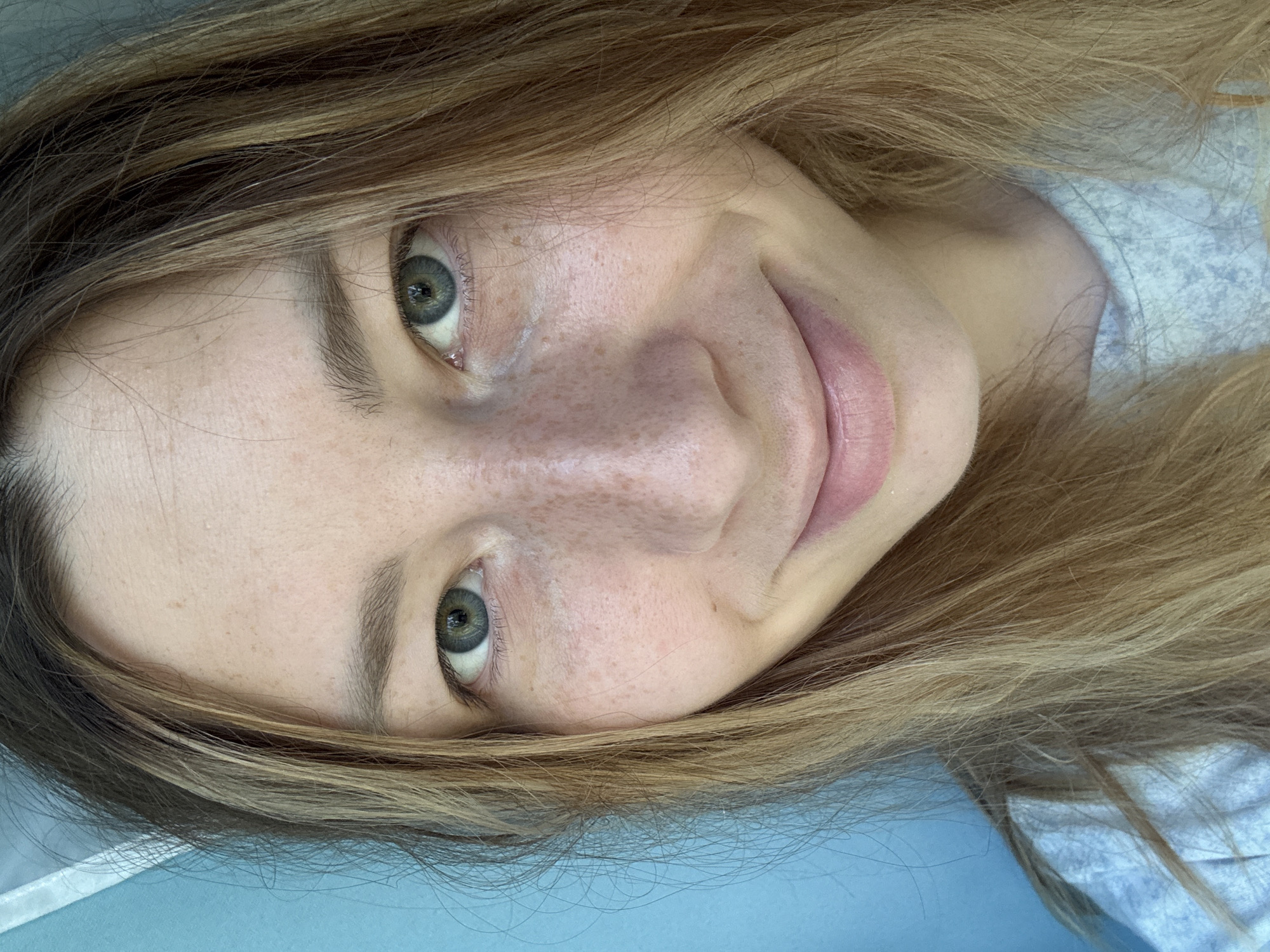 A close-up, rotated portrait shows a woman with light skin and freckles on her nose and cheeks. She has light brown hair with blonde highlights, visible around her face and extending to the edge of the frame. Her eyes are greenish-gray. She is smiling slightly. The background consists of light blue and white fabric, potentially a sheet or clothing. There is no information to suggest this image was taken in Moscow, Russia.